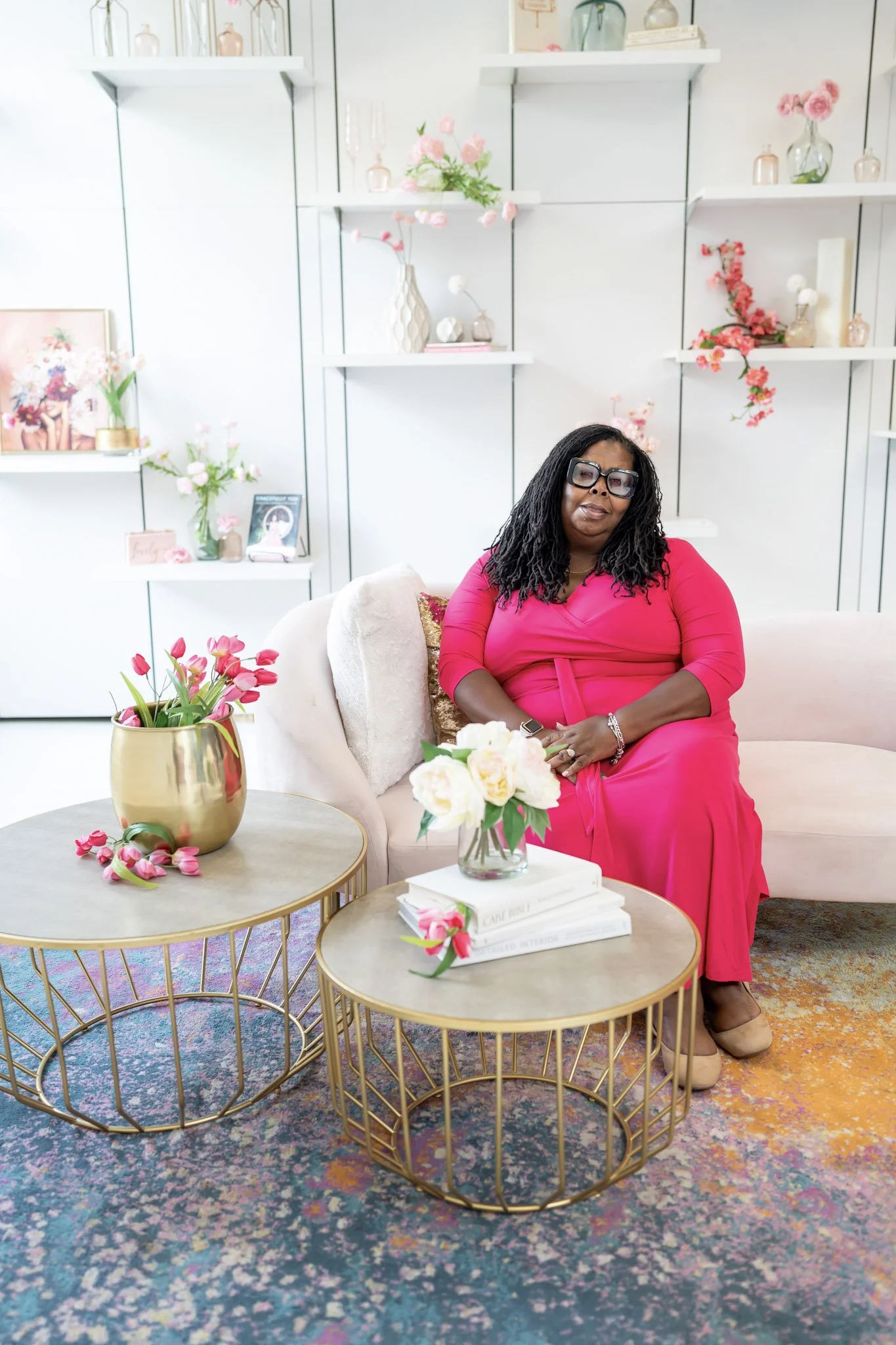 A woman wearing a bright pink dress and glasses sitting on a light-colored sofa with pink and white flowers on tables and shelves behind her in a decoratively styled room.