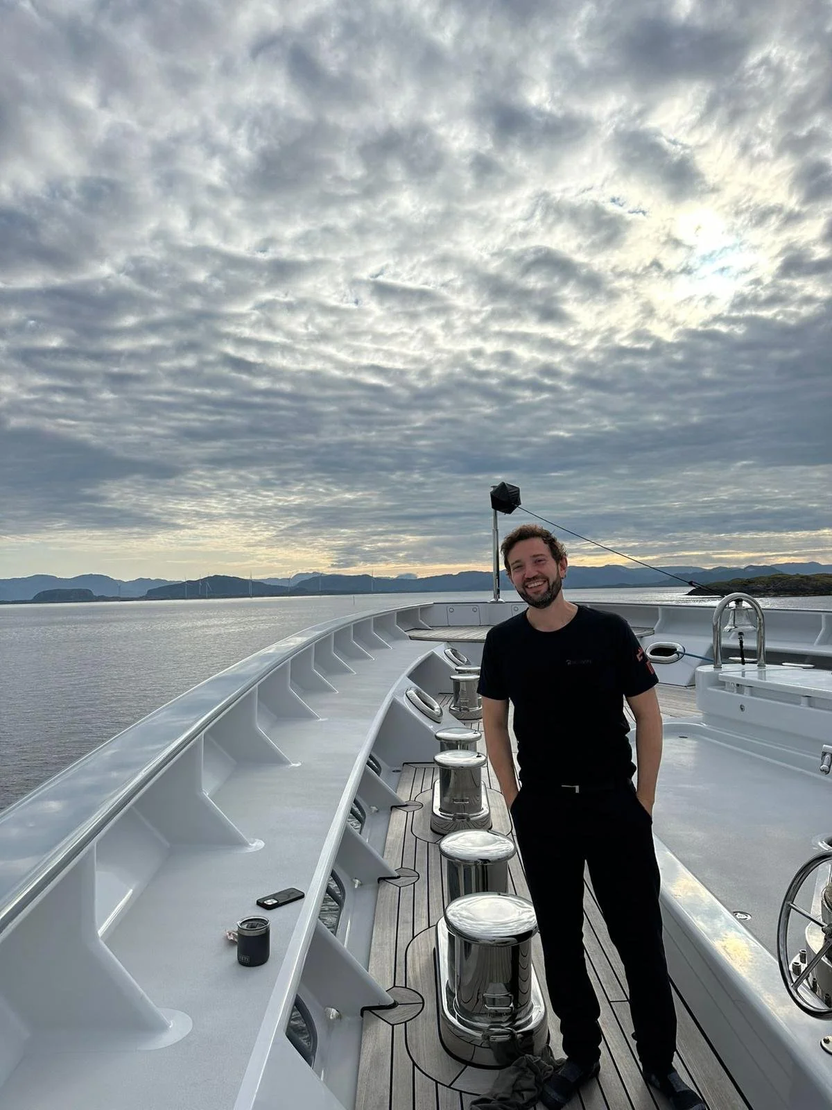 A smiling man in black clothes standing on the deck of a boat with water and mountains in the background under a cloudy sky.