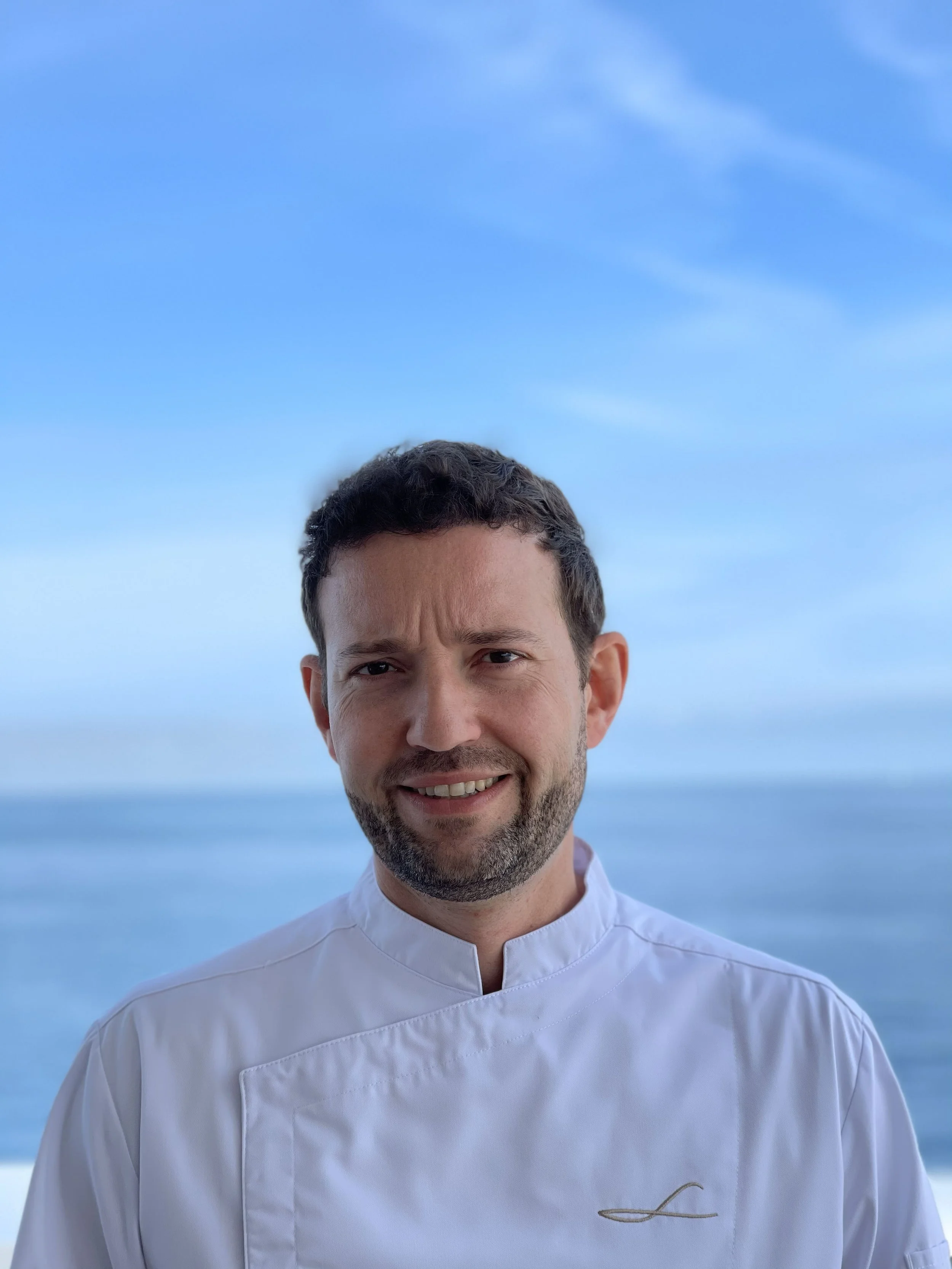 A man dressed as a chef, smiling, with an ocean and blue sky in the background.