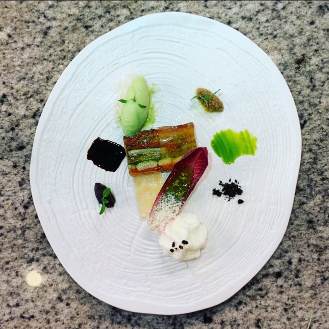 Elegant plated food with various colorful, artfully arranged bites including a dollop of white cream, a slice of cured meat, green vegetables, and garnishes on a textured white plate.