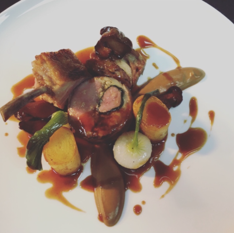 A plated gourmet dish featuring a piece of meat wrapped in a cut of bone, garnished with small vegetables including baby onions and green onions, with a dark sauce drizzled artistically around the plate.