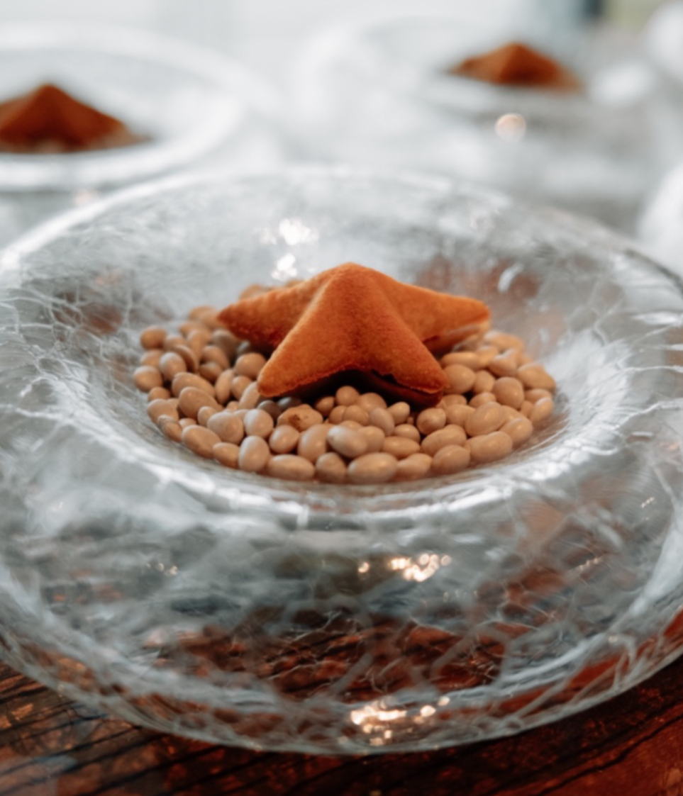 A glass bowl containing beige beans and two triangular, brown, fried snacks on top. The bowl is placed on a wooden surface.