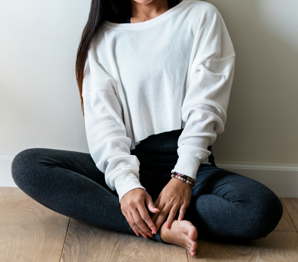 Woman sitting cross-legged on the floor in a white long-sleeve shirt and dark leggings, with her hands resting on her ankle, near a white wall.