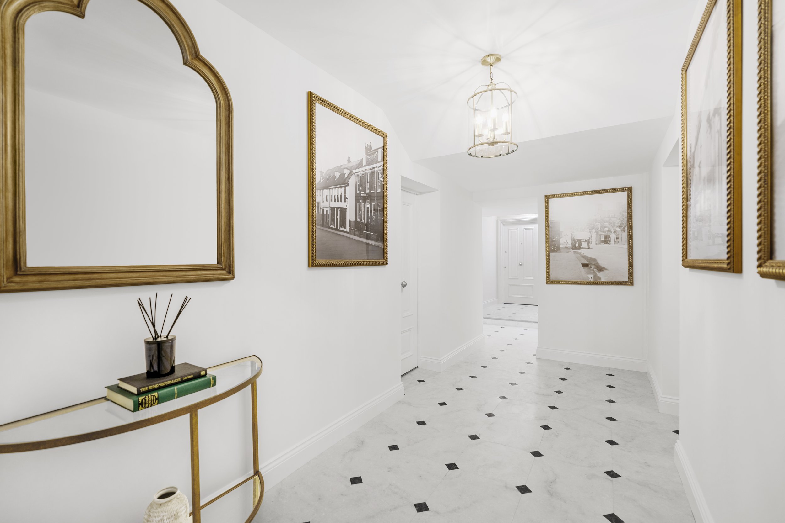 Bright white hallway with framed black-and-white photographs, a gold-framed mirror, a gold and white console table with books and a reed diffuser, white walls, and a chandelier.