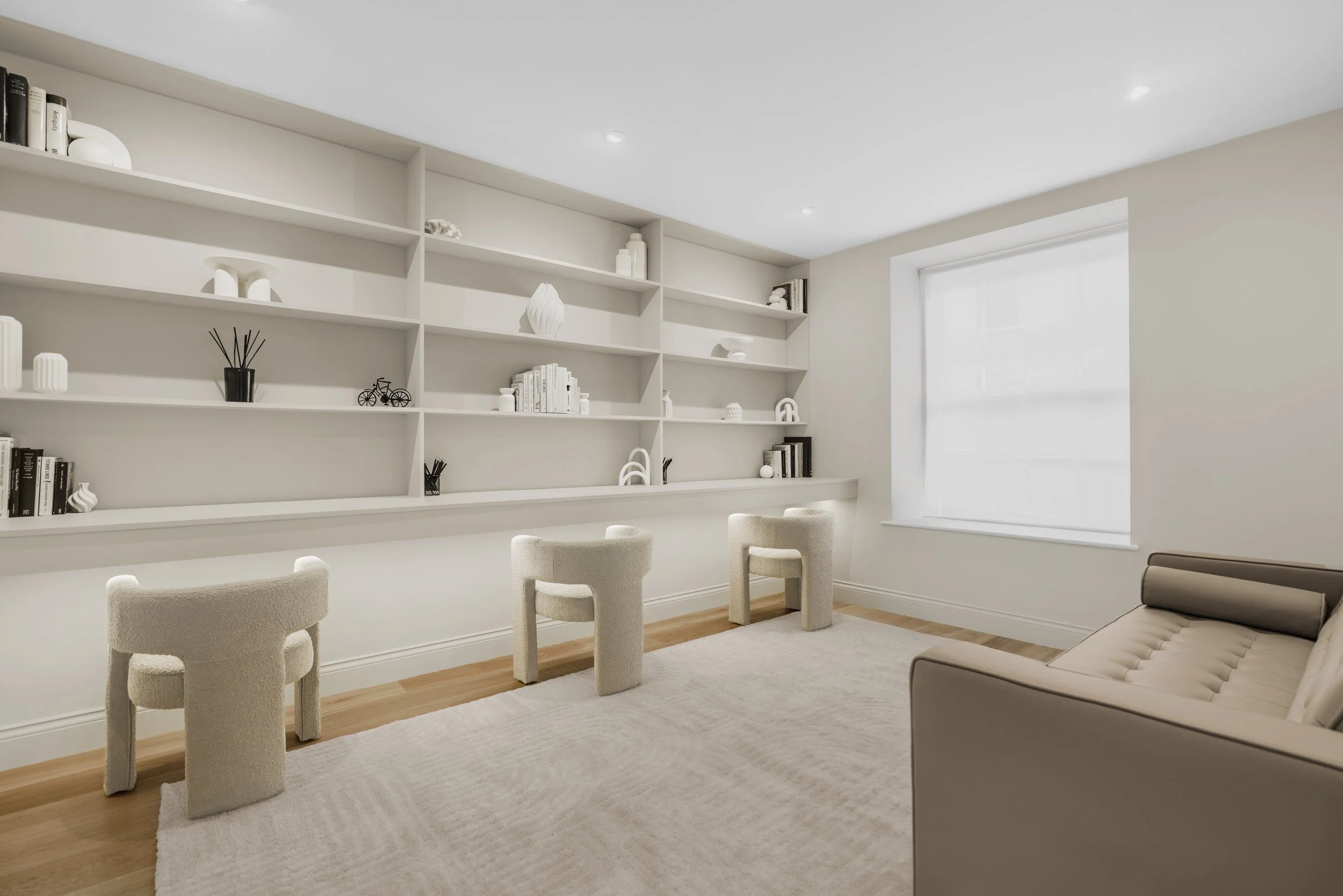 Minimalist living room with built-in white shelving, cream armchairs, a beige sofa, and a large window with white blinds, decorated with white and black decorative items.