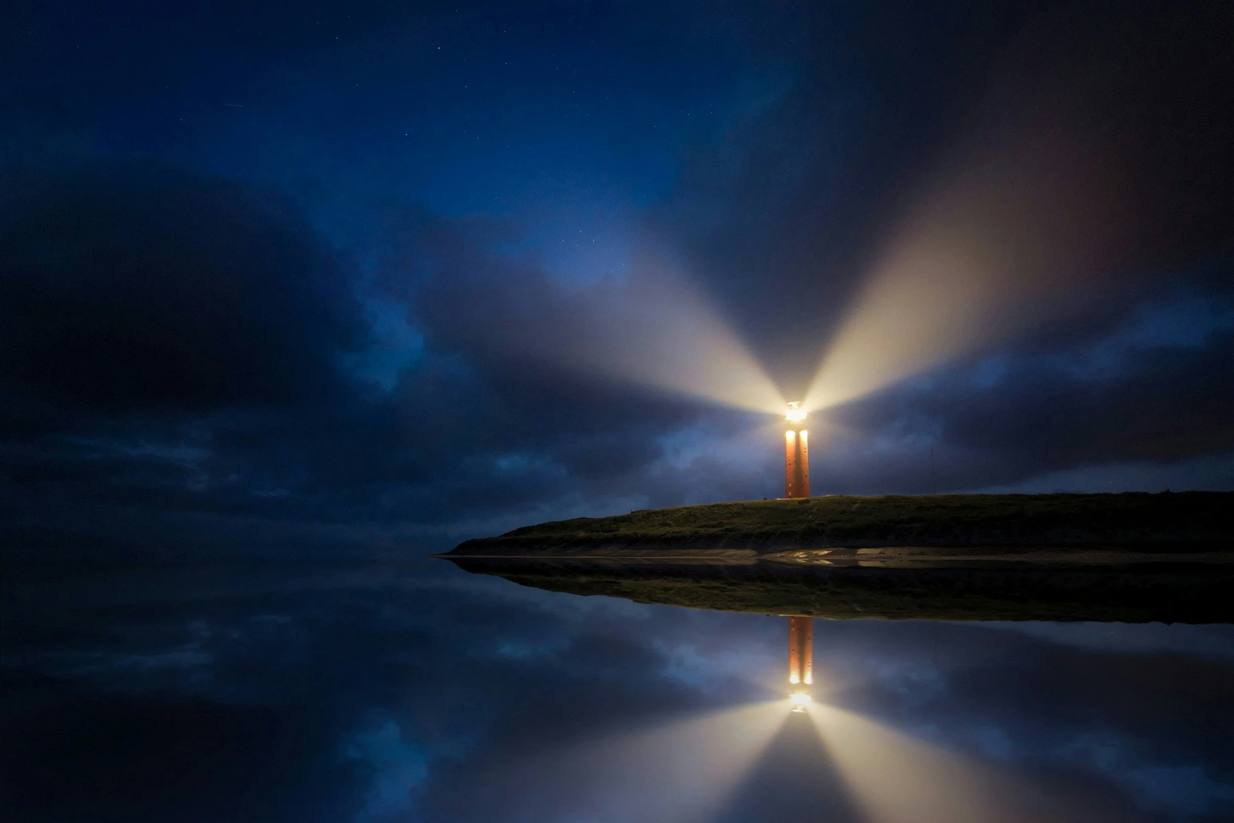 Lighthouse at night illuminating the dark, cloudy sky with its beam of light, reflected in calm water below.