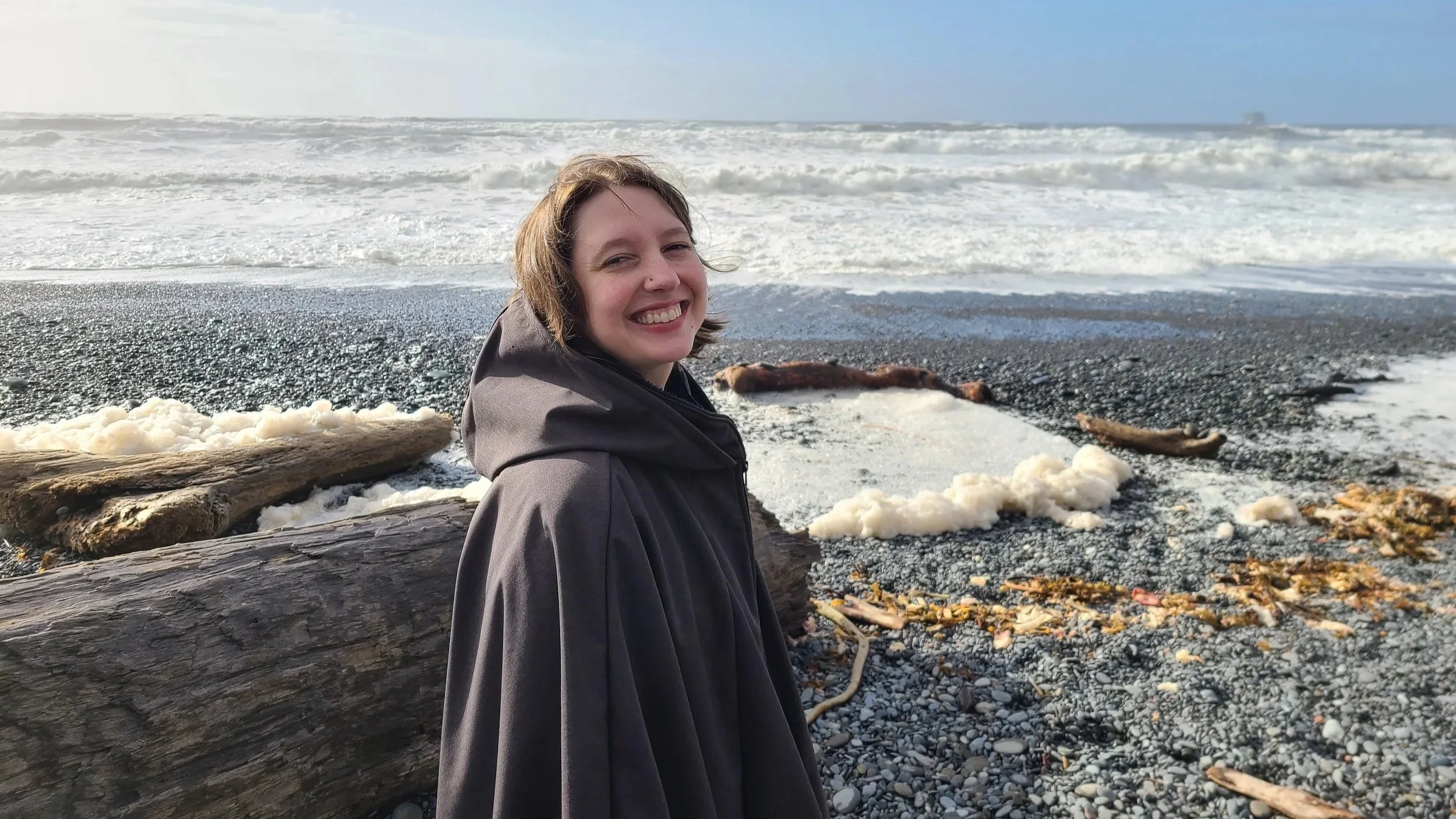 A white woman with chin-length hair, wearing a grey cape, smiling at the camera on a rocky beach near the ocean with waves and driftwood in the background.