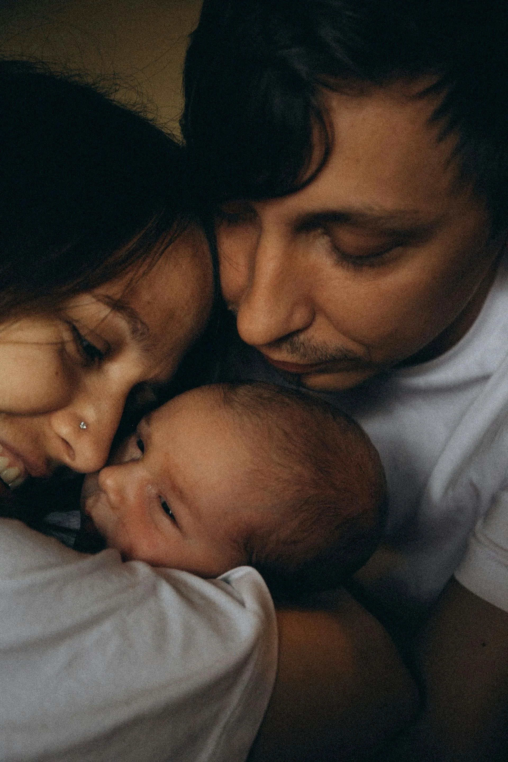 A family of three, a man, a woman, and a baby, close together with their faces touching and smiling, engaging in a tender and joyful moment.
