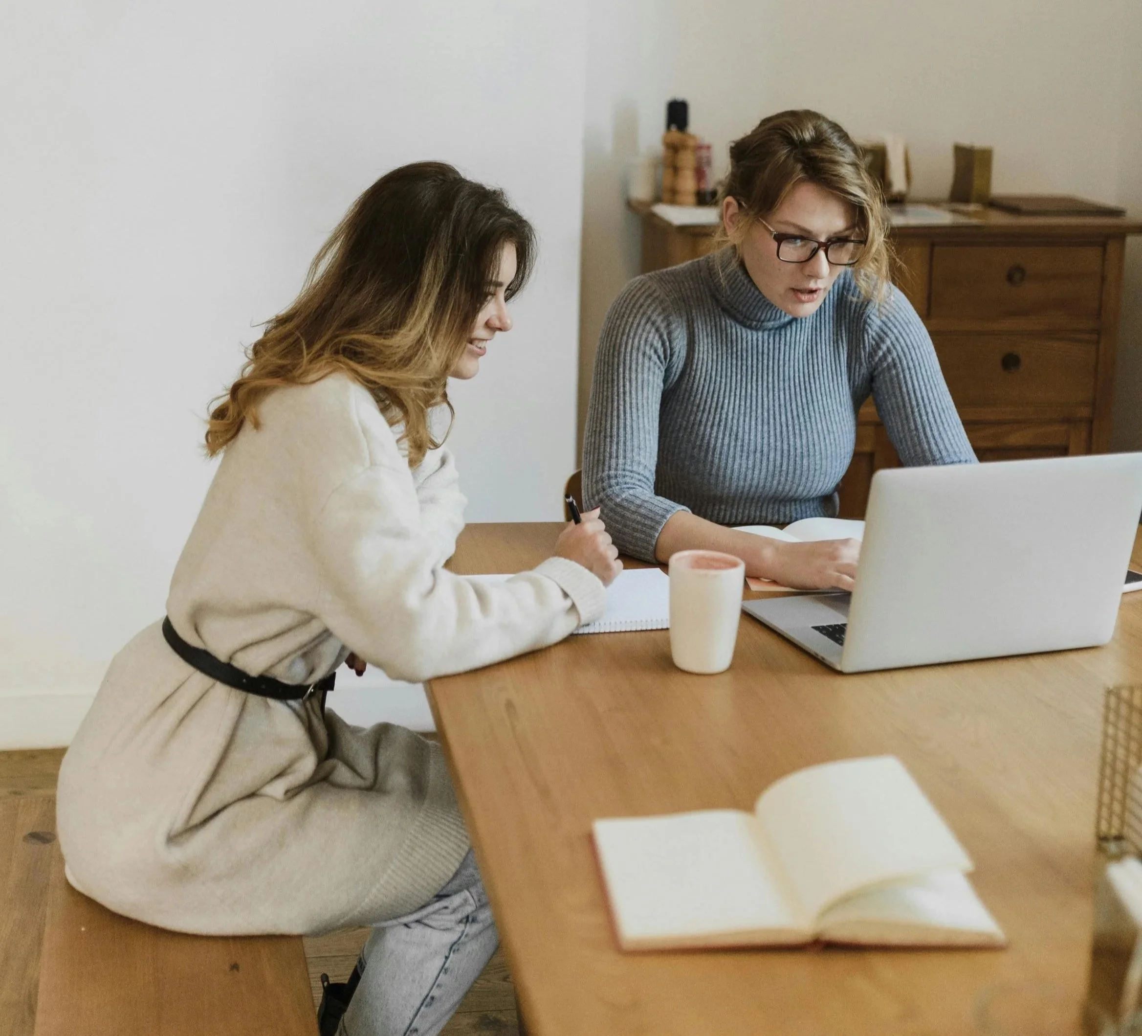 Two women sitting at a wooden table working together. One is writing in a notebook, and the other is working on a laptop. There are notebooks, a candle, and an open book on the table.