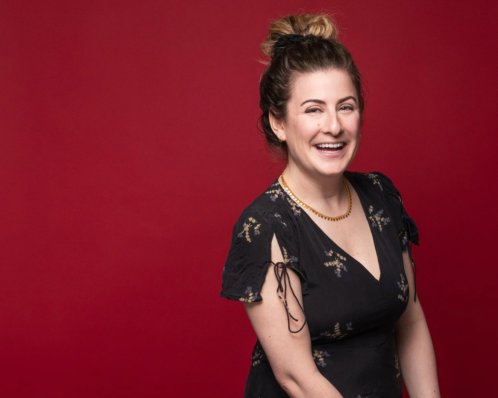 A woman with brown hair in a messy bun, wearing a black dress with lace-up sleeves and a gold necklace, smiling in front of a red background.