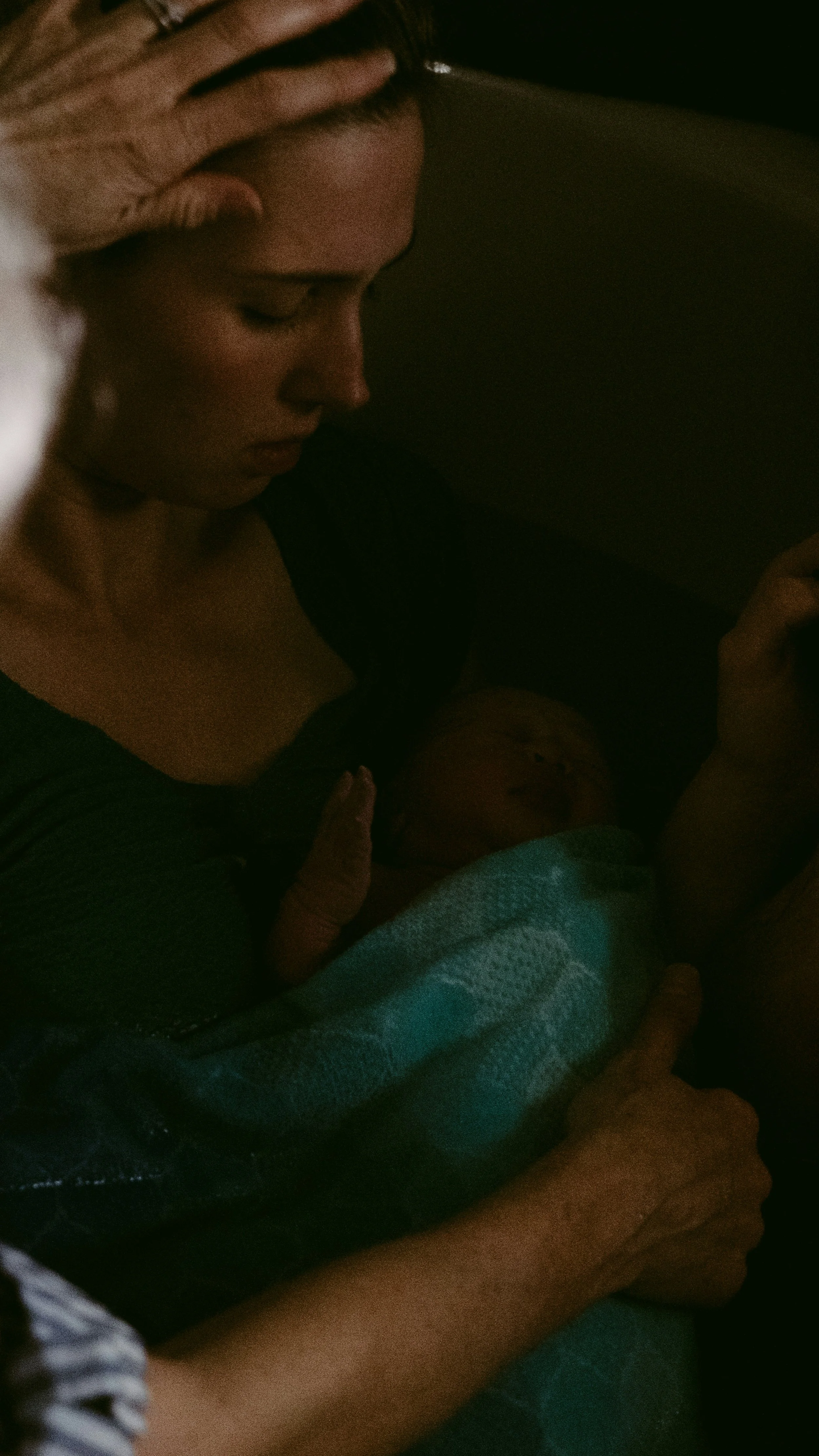 A woman with closed eyes rests her head on her hand as she holds a newborn baby wrapped in a blanket, lying on her chest.