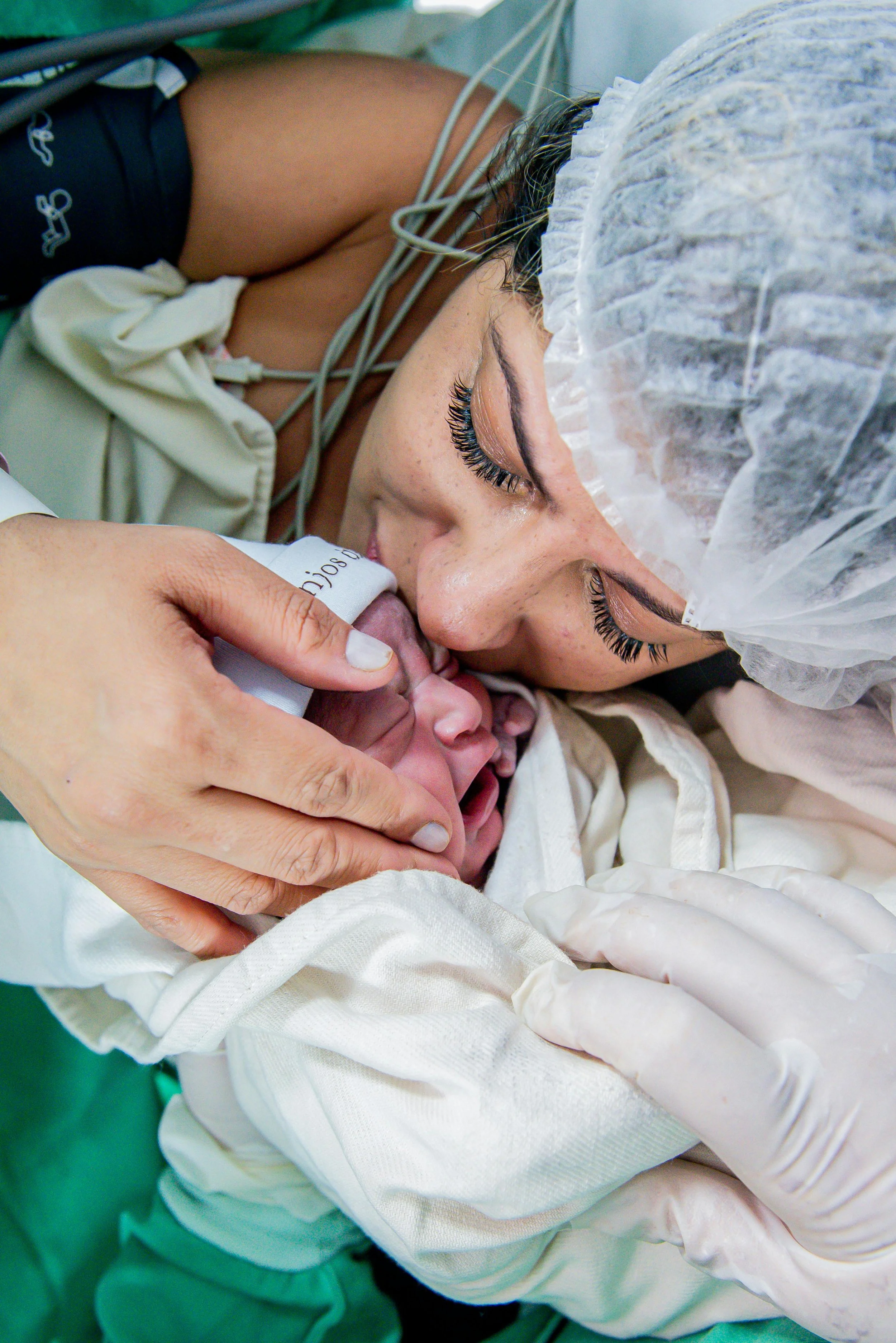 A woman with long eyelashes and a hair cover kisses a newborn baby during birth at a hospital, with medical tools and gloves seen.