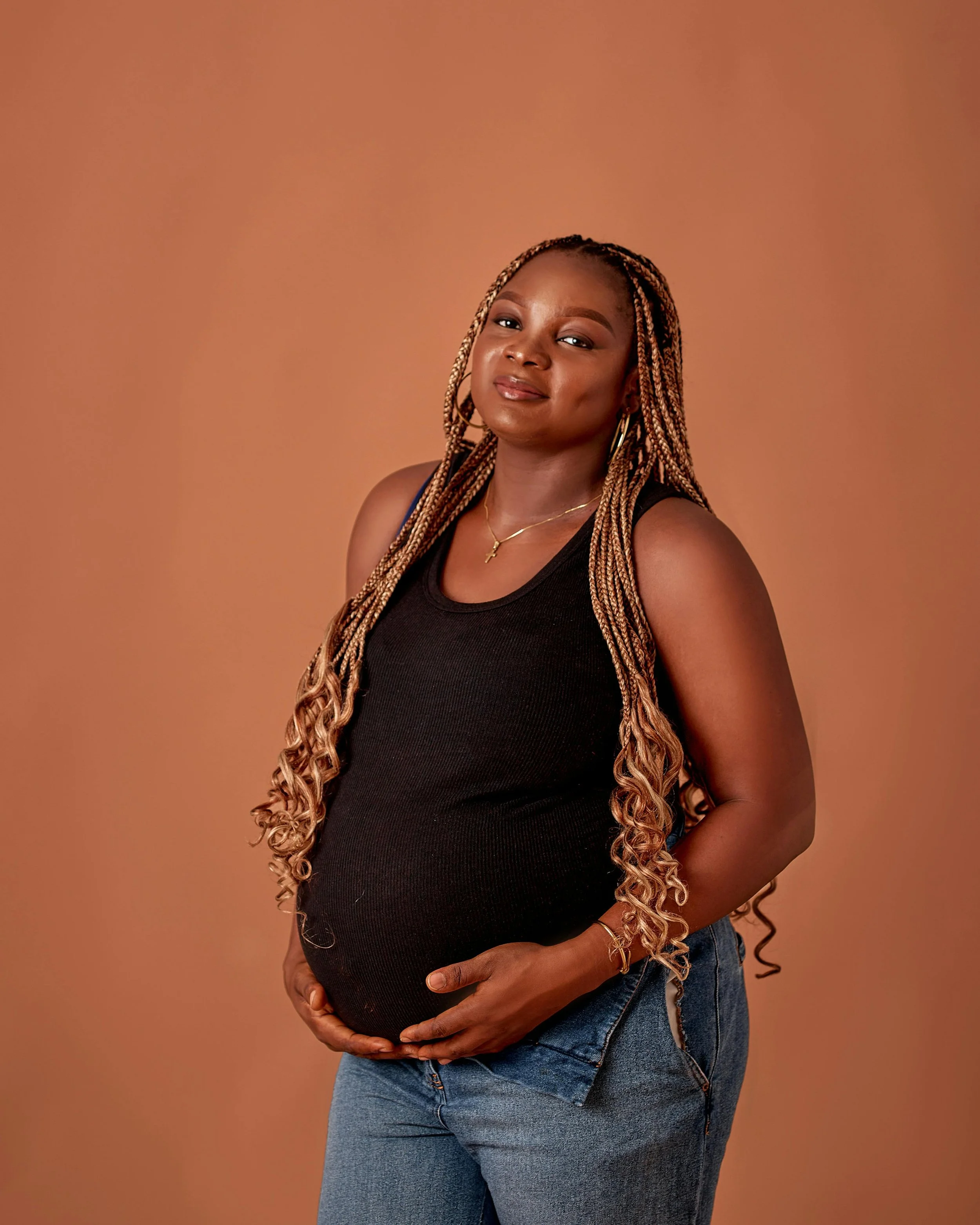 Pregnant woman with long curly braids wearing a black tank top and jeans, standing against a beige background.