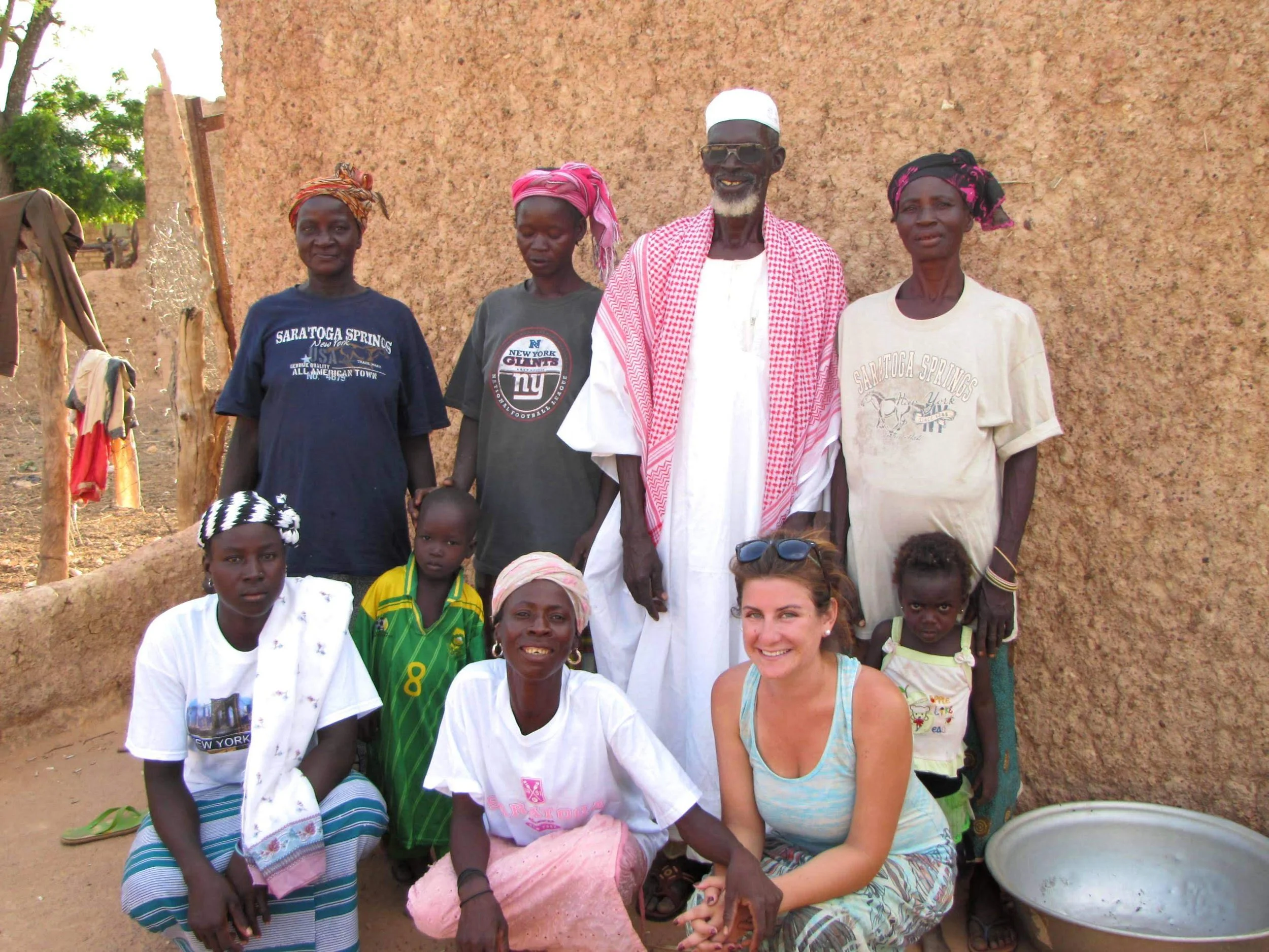 A group of people, including women and girls, gathered outdoors in front of a mud wall, some wearing colorful headscarves and casual clothing, with a woman and a man smiling among them.