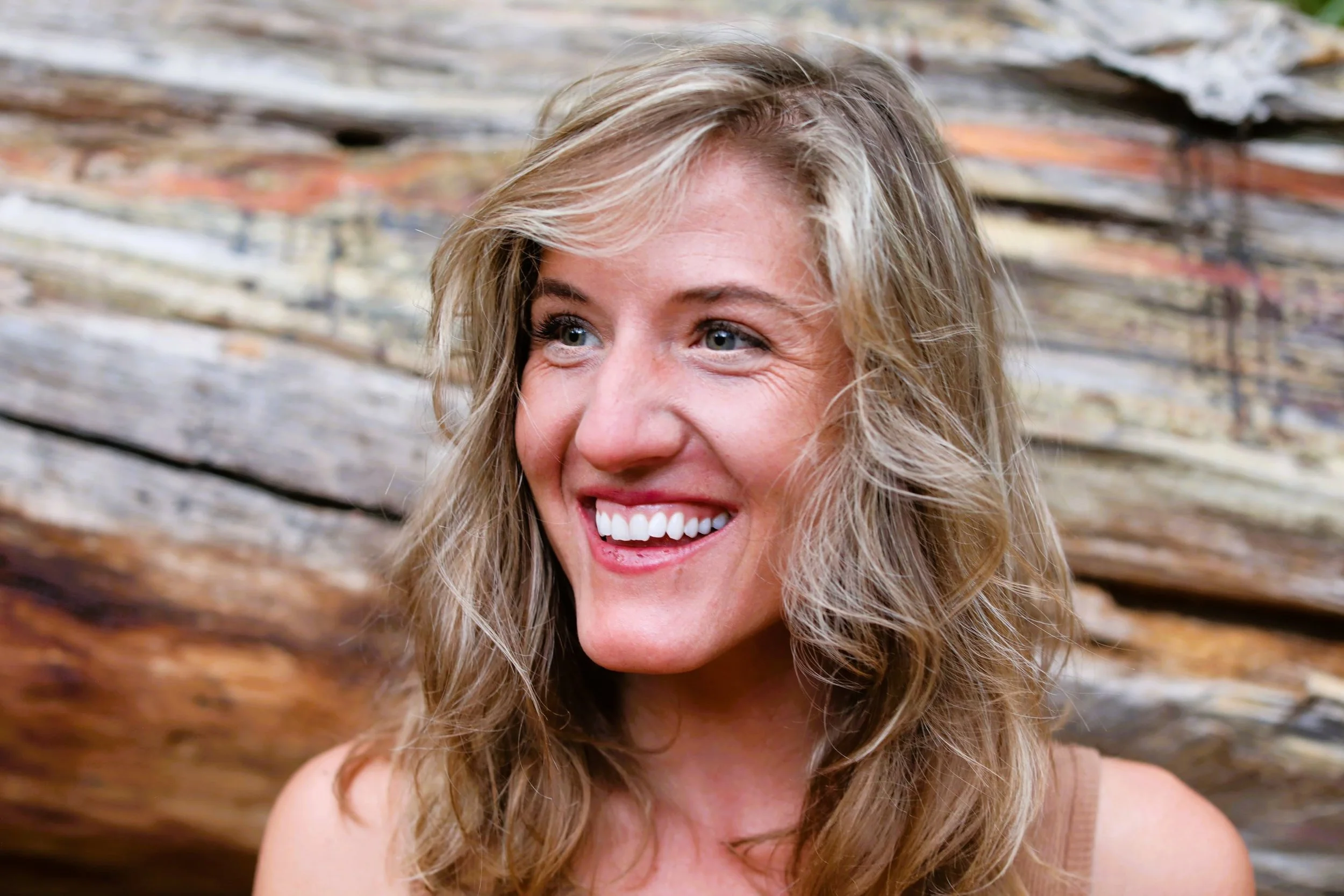 A smiling woman with wavy blonde hair and blue eyes, outdoors in front of a background of weathered wood logs.