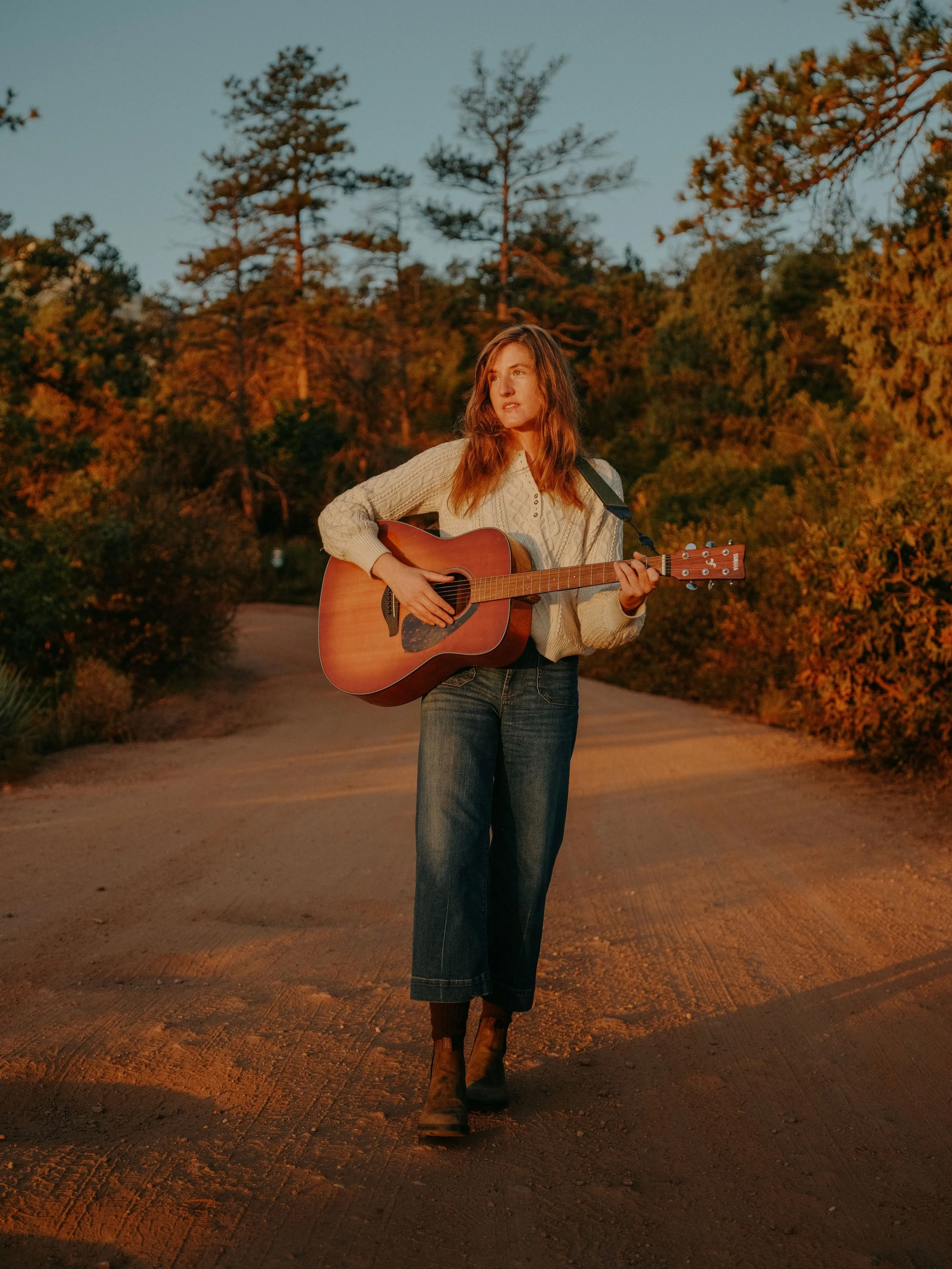 A young woman walking on a dirt road playing an acoustic guitar during sunset, surrounded by trees with autumn foliage.