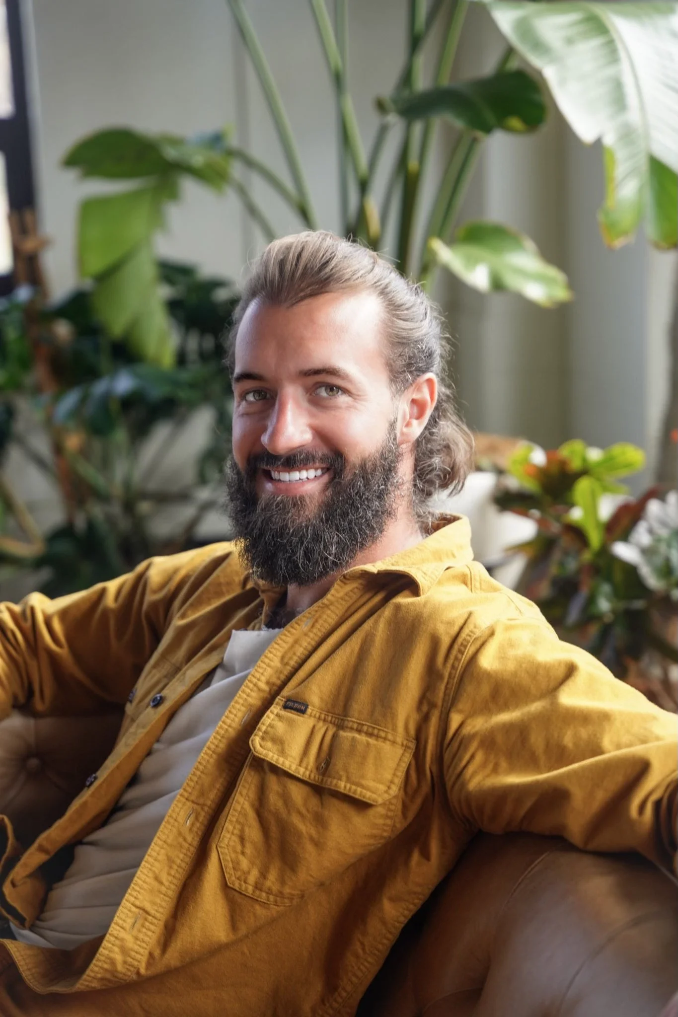 A man with a beard and long hair, smiling, sitting on a couch in front of green plants.