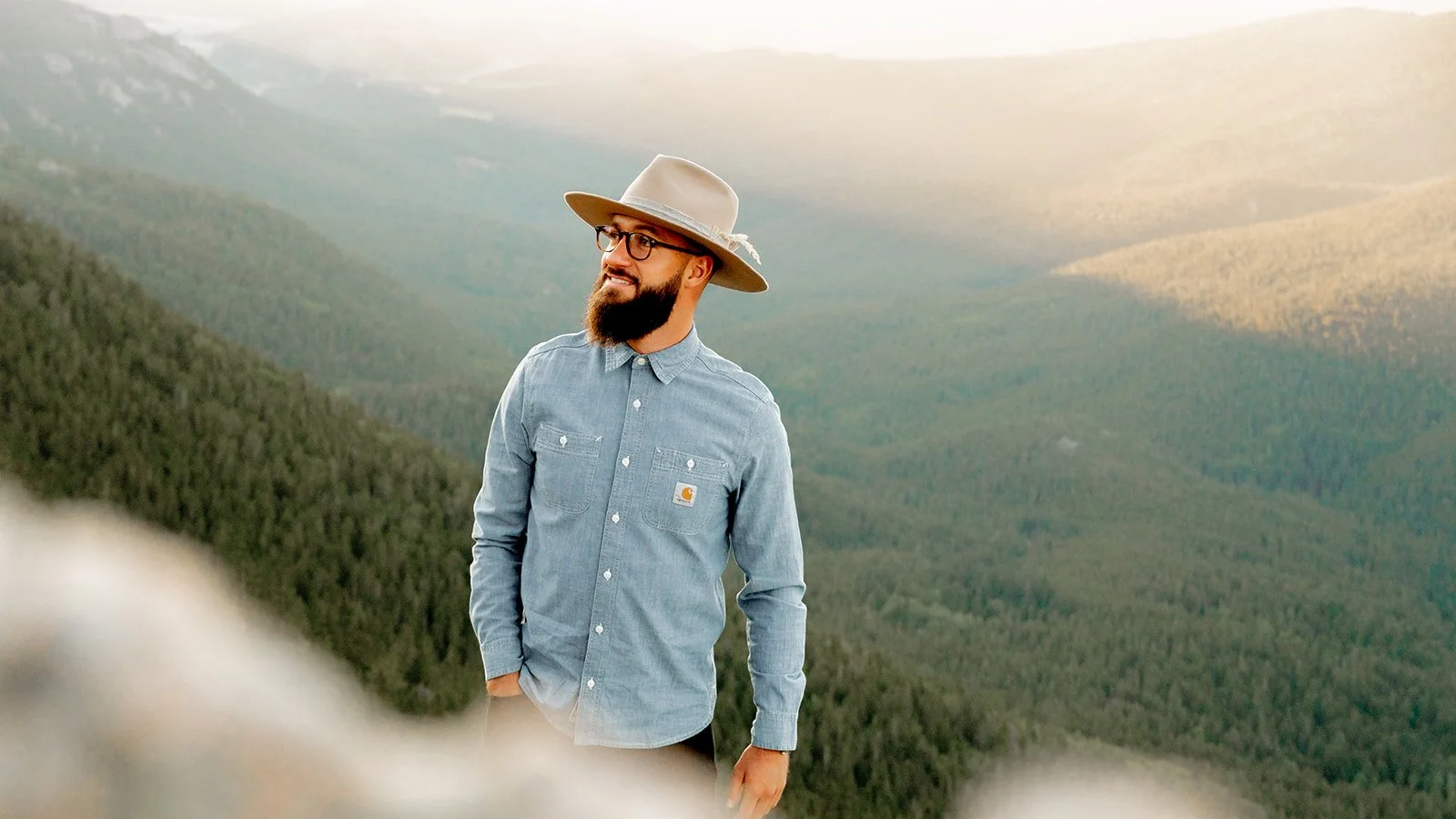 A man with glasses and a beard wearing a wide-brimmed hat and denim shirt standing outdoors on a mountain with a forested landscape.