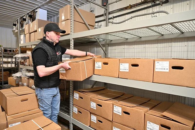 A man in a black cap and vest is placing a cardboard box on a warehouse shelf filled with similar boxes, in a storage or distribution facility.