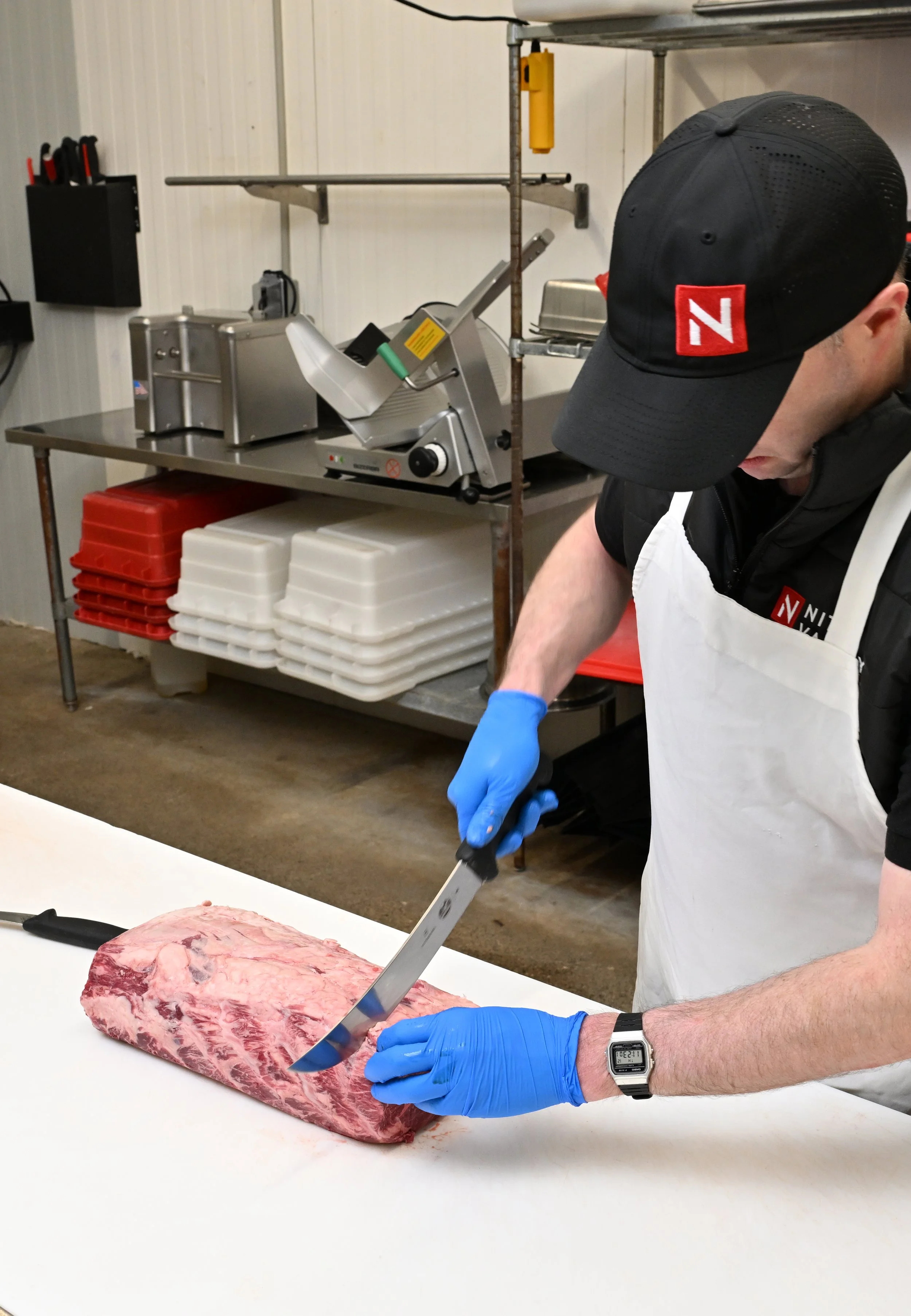 A person wearing a black cap with a red and white logo, blue gloves, and a white apron is slicing a large piece of raw meat on a white cutting board in a kitchen or meat processing area.