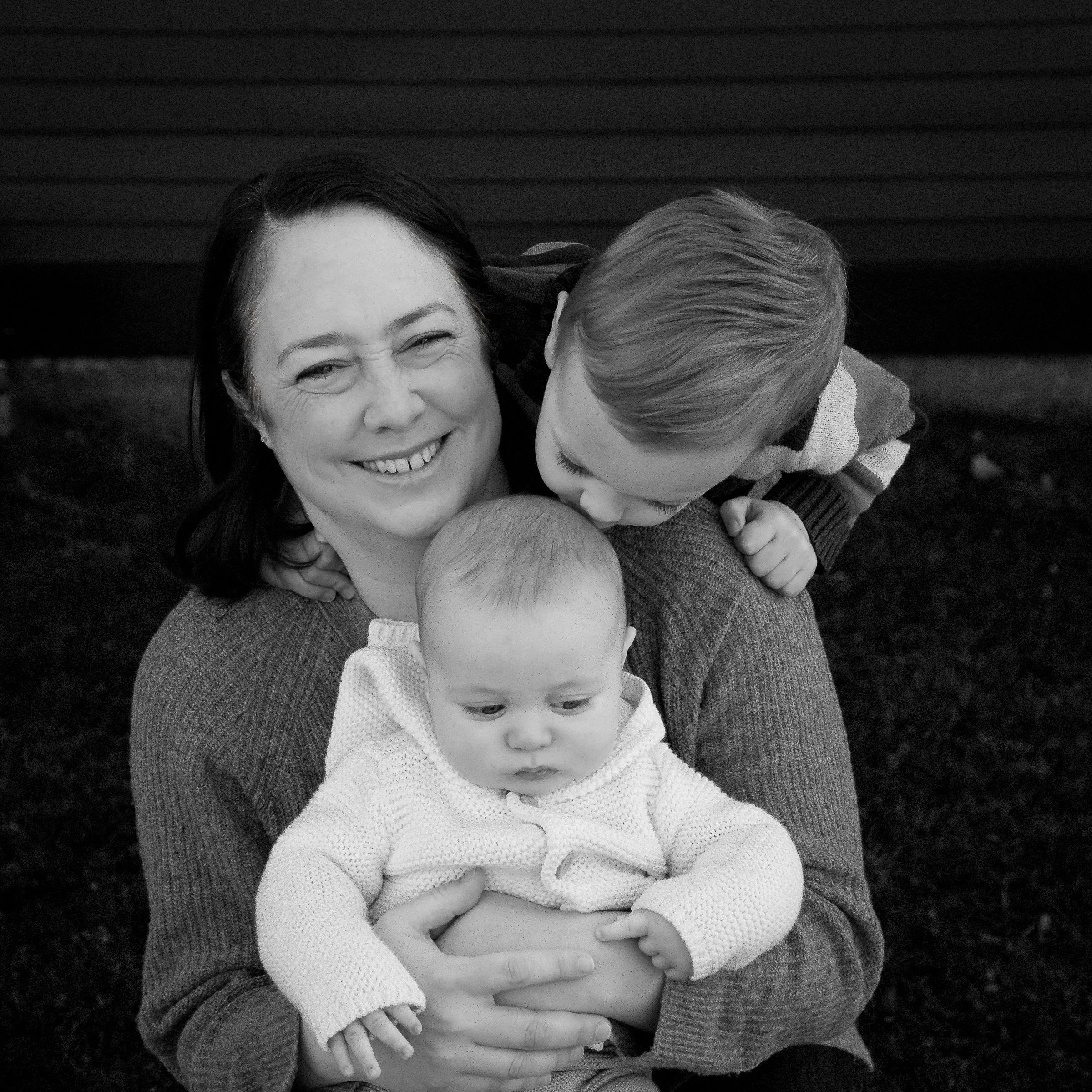A woman smiling and holding two young children, one child kissing her cheek, all outdoors.
