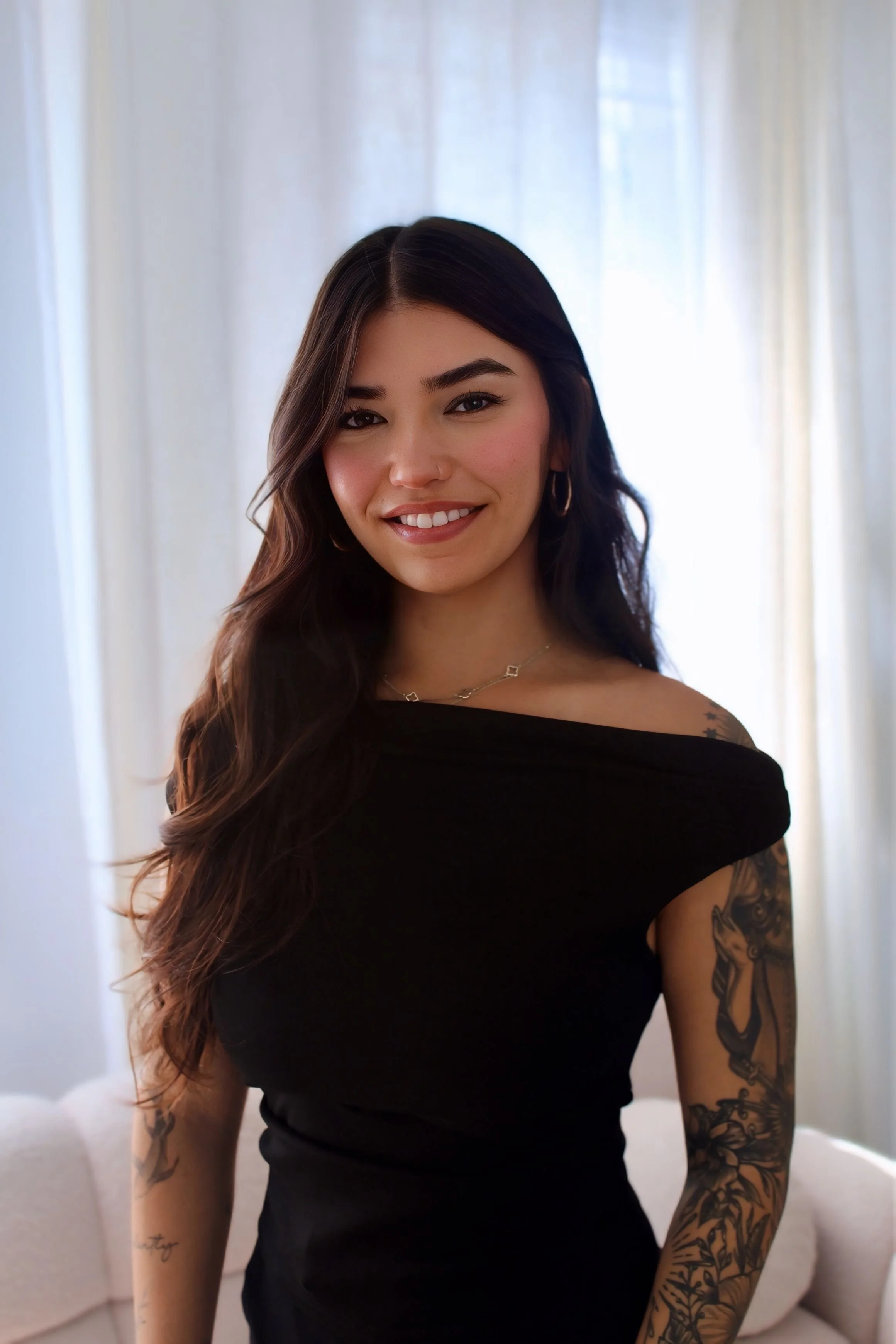 A young woman with long dark hair, wearing a black off-shoulder top, smiling at the camera in a well-lit room with curtains and a sofa in the background.