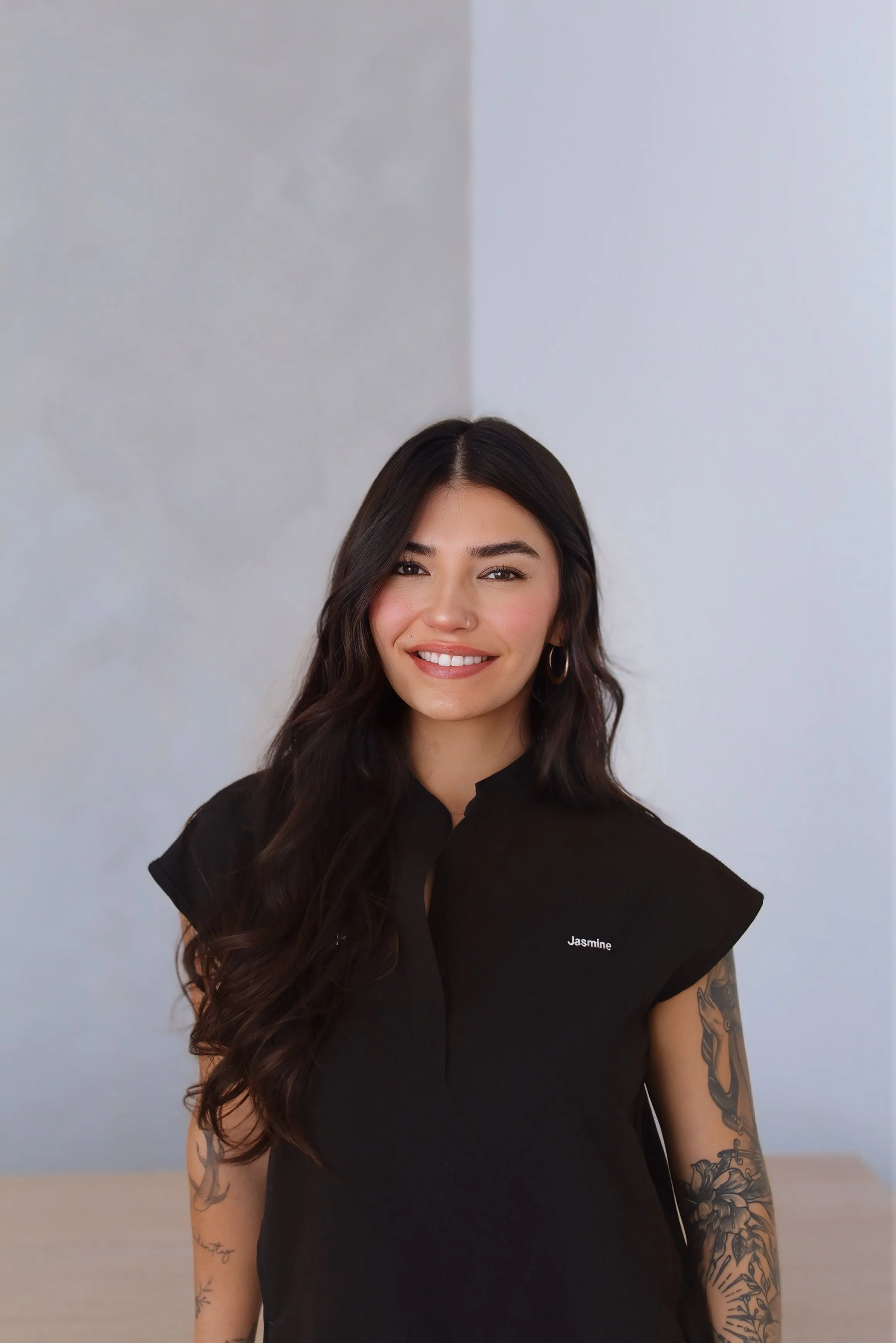 A smiling woman with long dark hair, wearing a black shirt with the name "Jasmine" embroidered on it, standing indoors against a plain background.