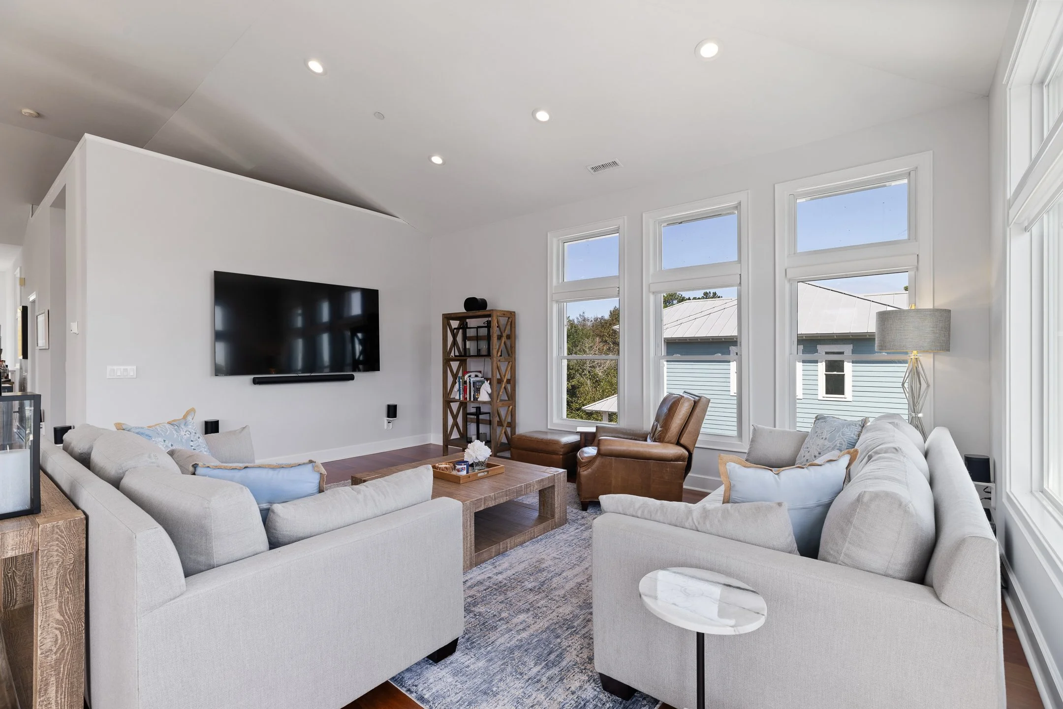 Living room with two beige sofas, a brown armchair with ottoman, a wooden coffee table, a large flat-screen TV on the white wall, a tall wooden bookshelf, a floor lamp, and three windows showing the exterior rooftops.