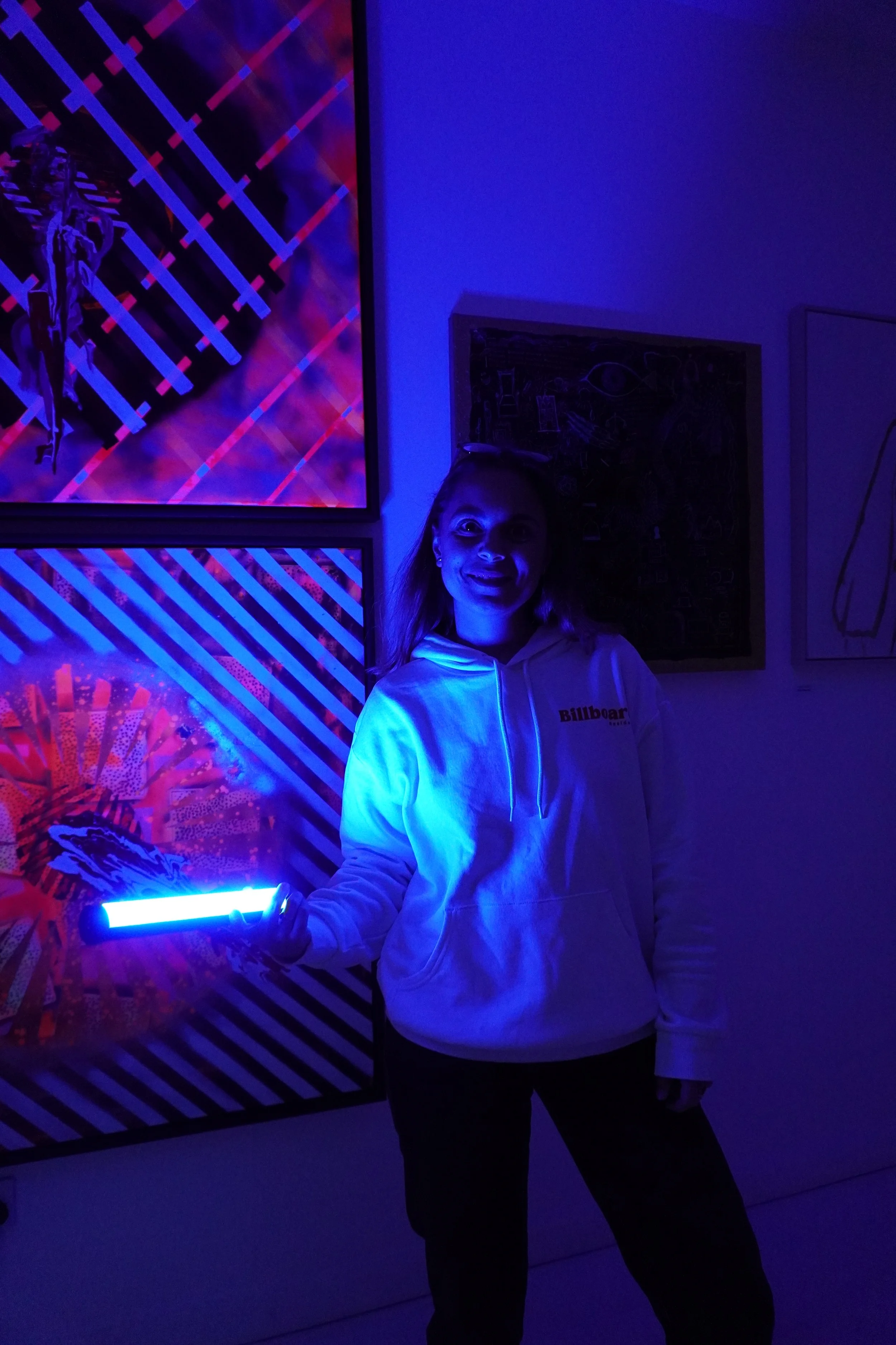 A young woman standing in a dimly lit room, holding a glowing blue light stick. She is smiling and wearing a white hoodie with political text on it. The room has vibrant, colorful artwork on the walls, illuminated by the glow of the light stick.