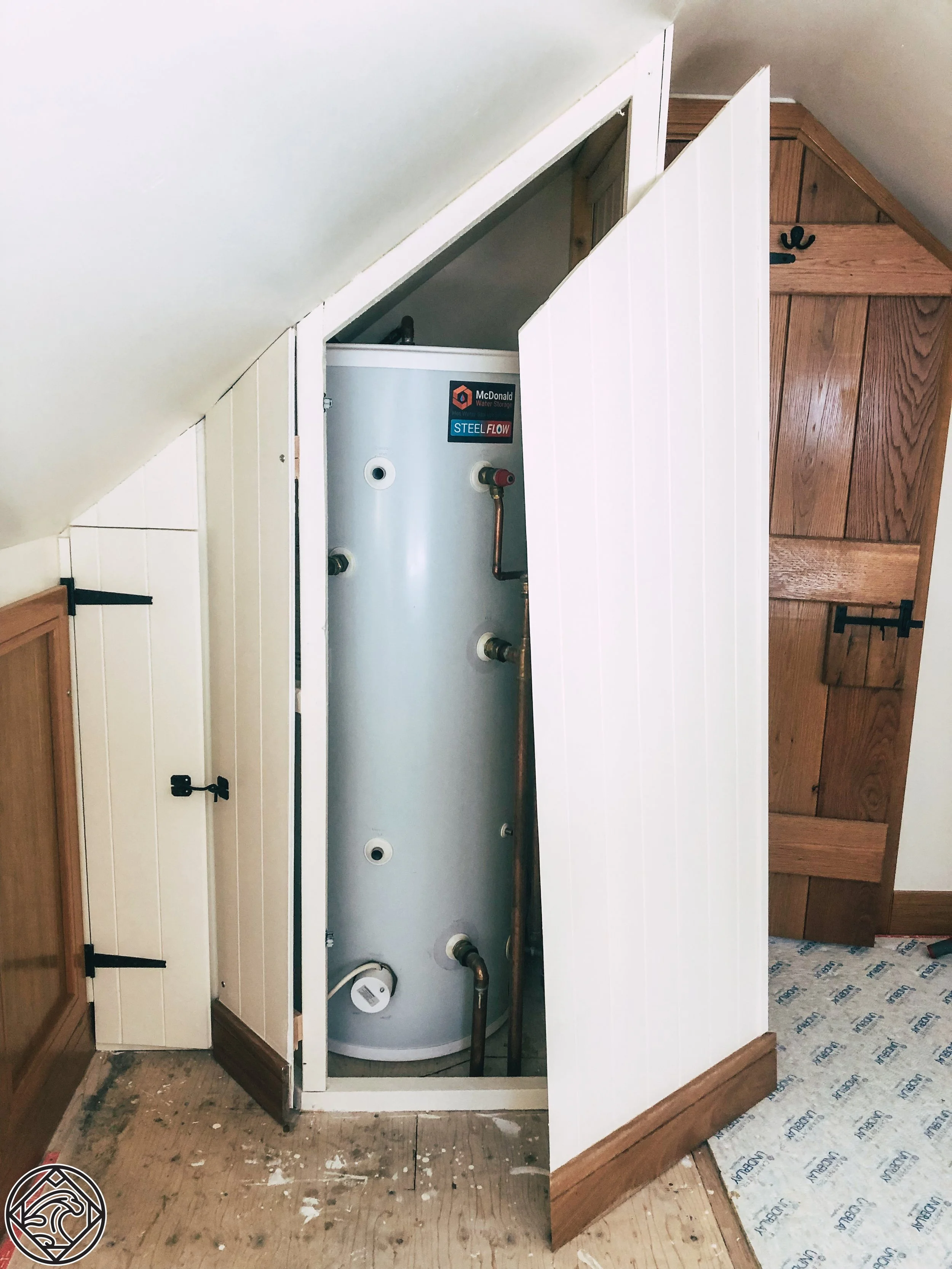 A water heater inside a wooden closet under a sloped ceiling, with white paneling and black hardware hinges, in a room with wooden flooring.