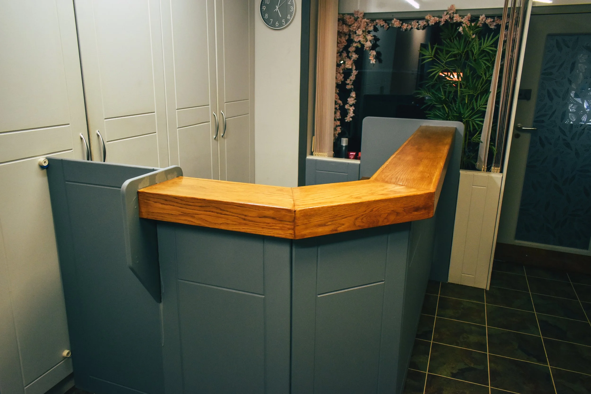 A reception desk with a wooden top and teal-colored base in a room with cabinet storage, a clock, and plants in the background.
