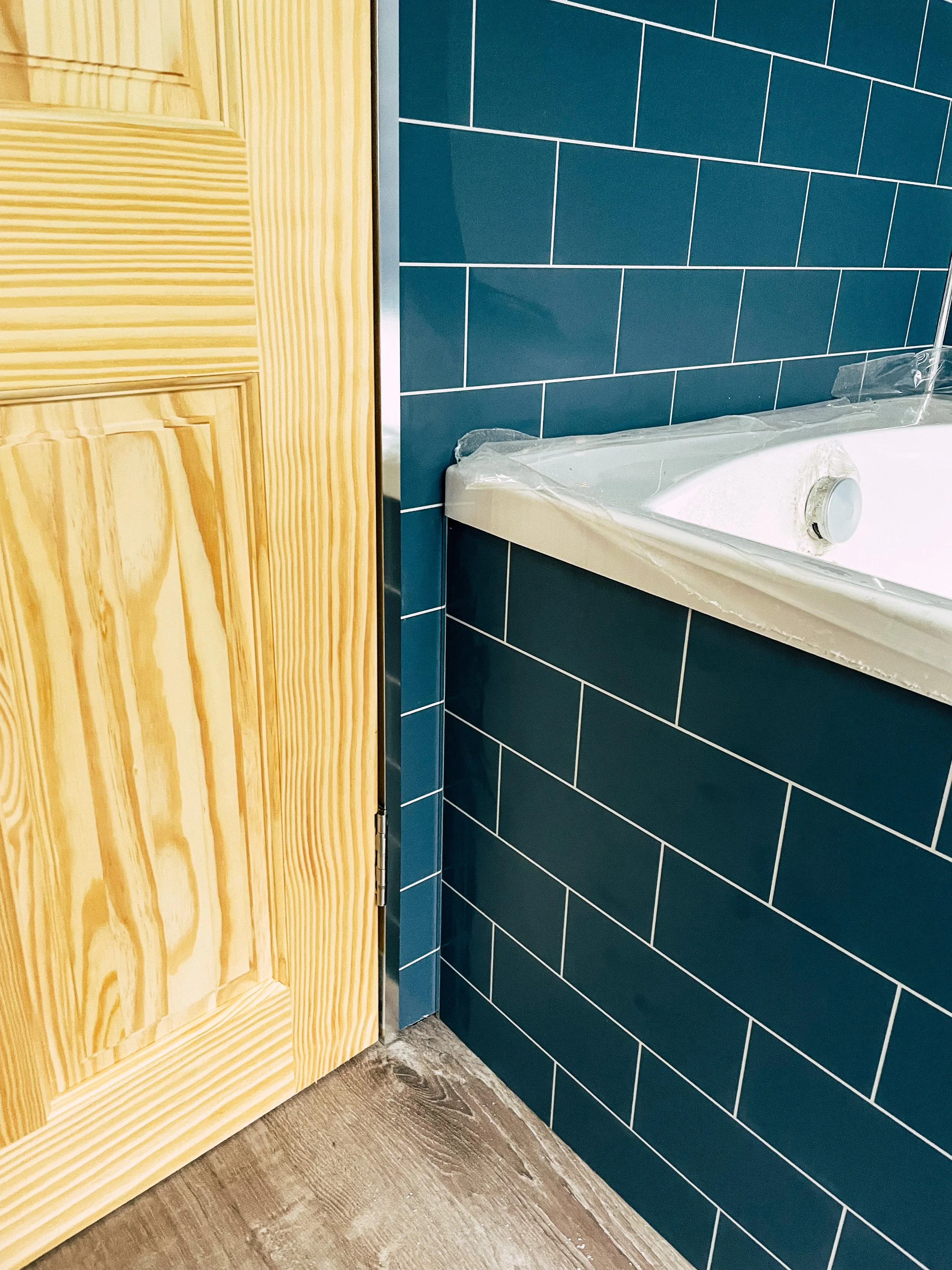 Close-up of a bathroom corner showing a blue tiled wall, a white bathtub with metal around the edge, and a wooden door.