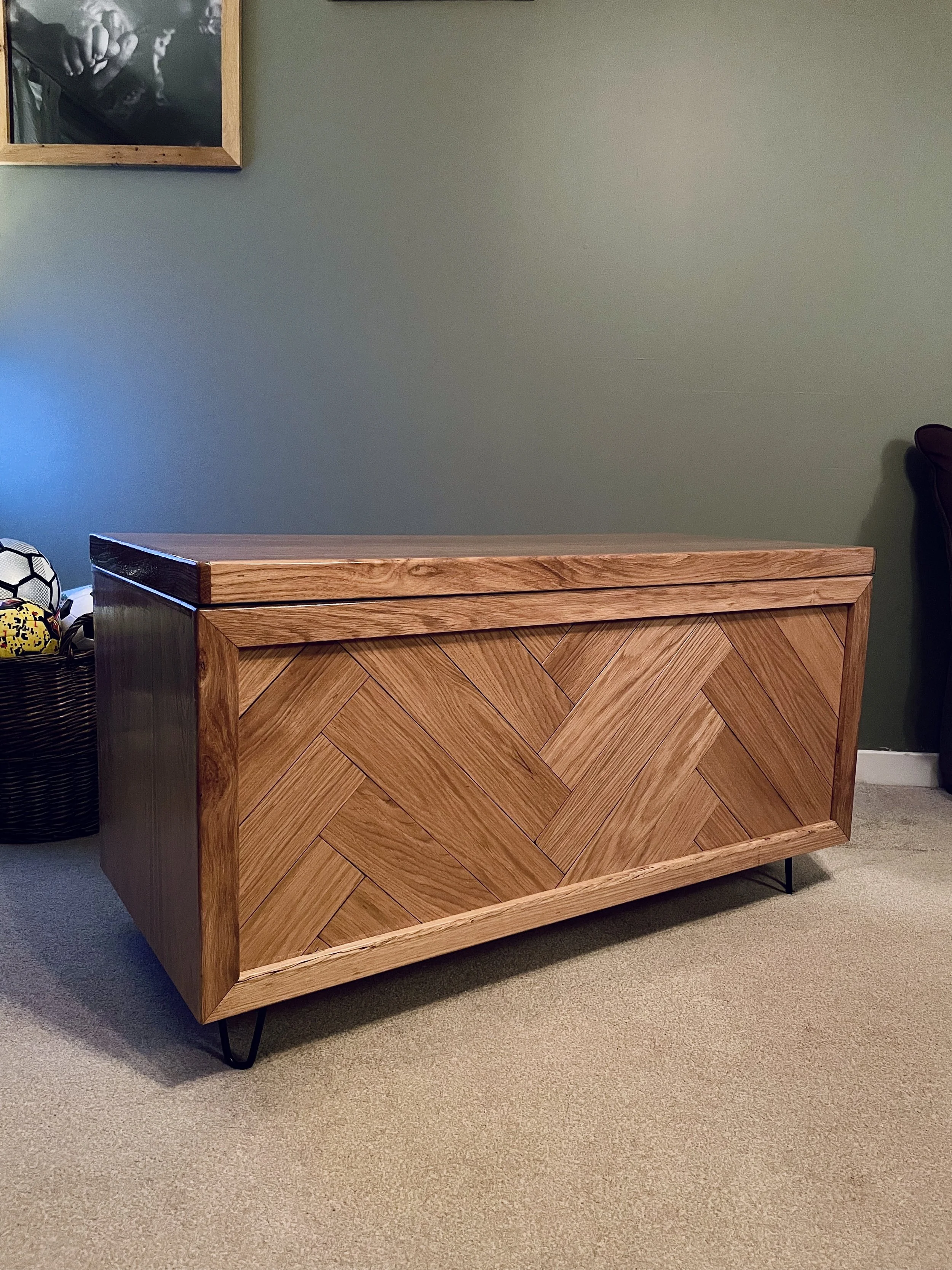 A wooden storage bench with a herringbone pattern on the front panel, standing on black metal hairpin legs in a room with green walls and beige carpet. 