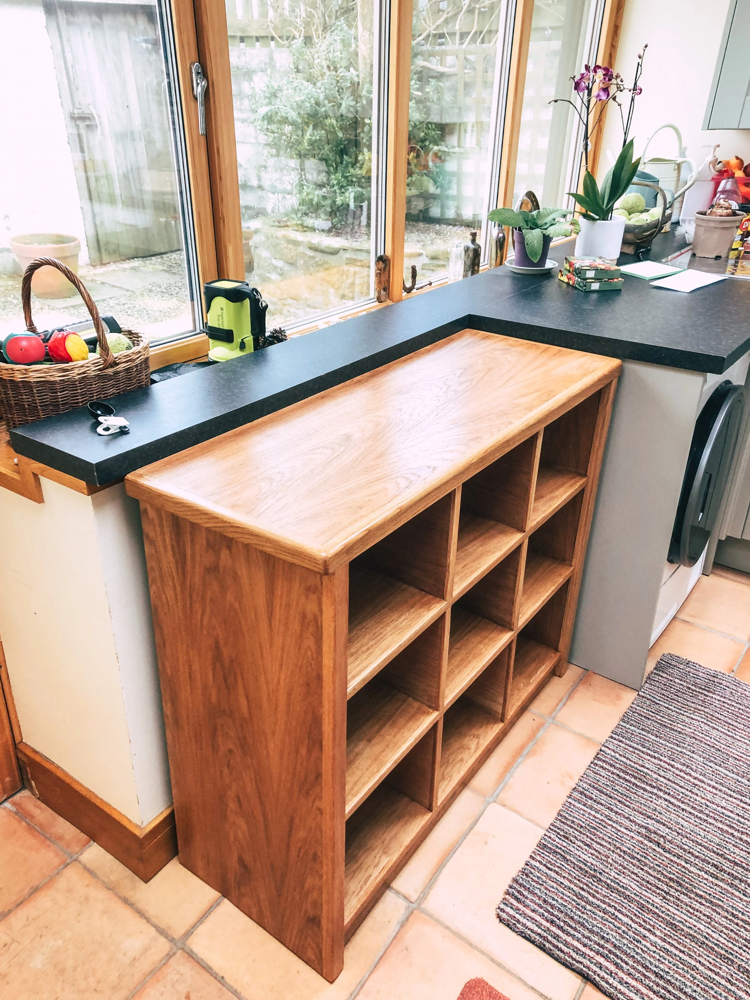 A wooden bookcase with nine open shelves placed against a kitchen counter near a window, with items such as a basket of toys, plants, and small miscellaneous objects on the counter.