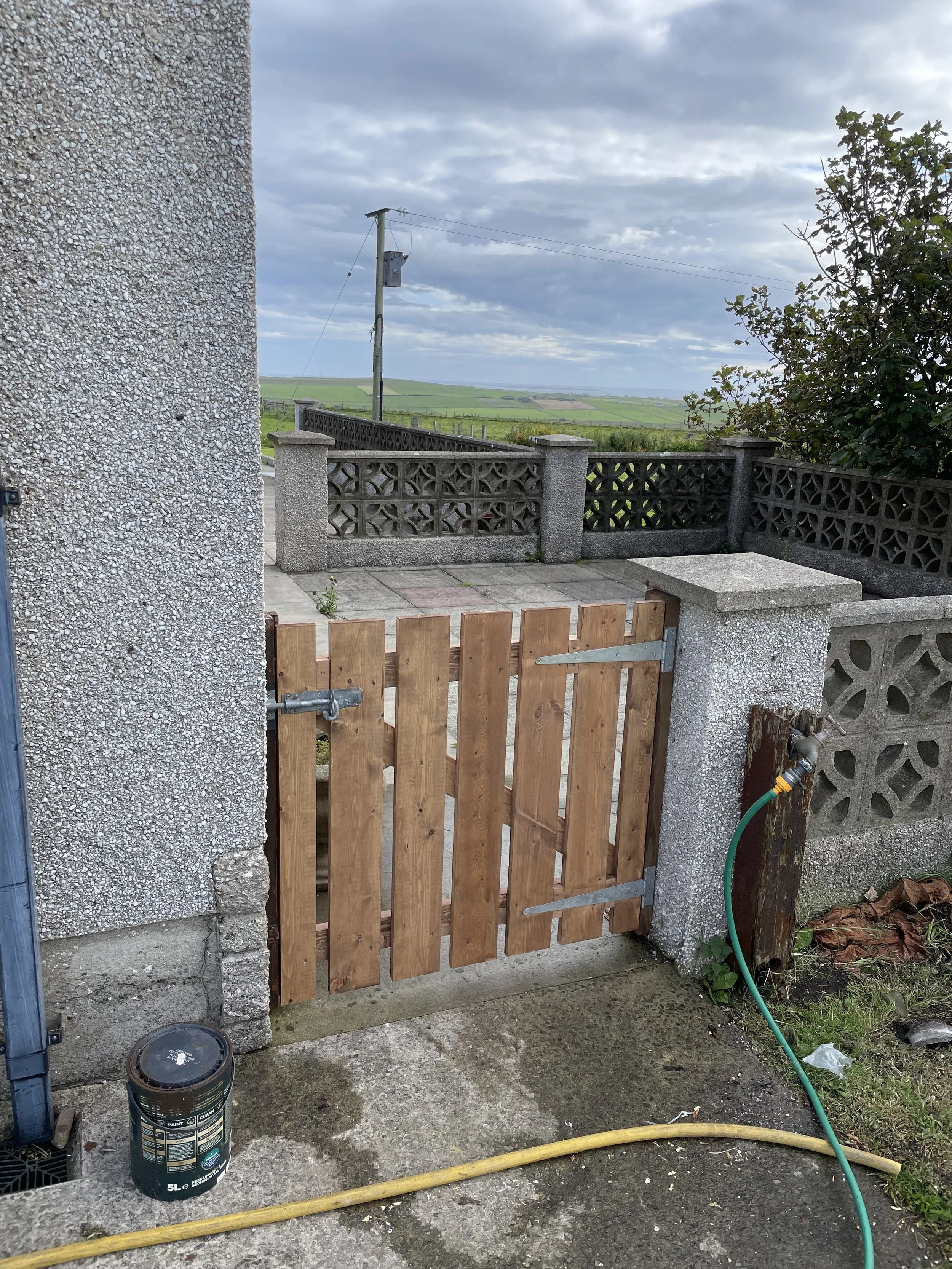 View of a small wooden gate installed in a concrete and stone wall, with a rain-soaked ground and a rural landscape with green fields and a cloudy sky in the background.