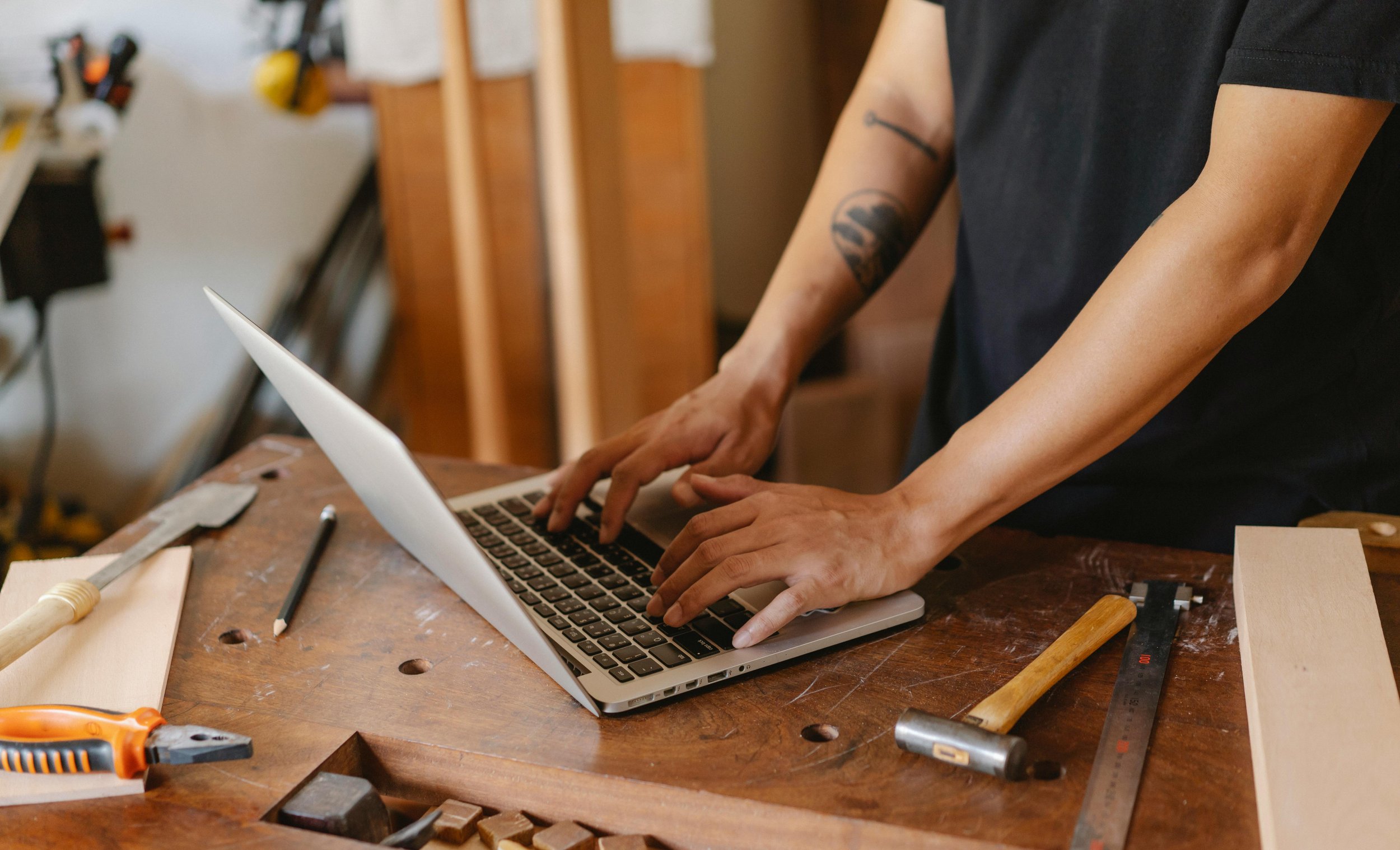 Person working on a laptop at a woodworking workshop, with tools like a hammer, chisel, and ruler on the workbench.