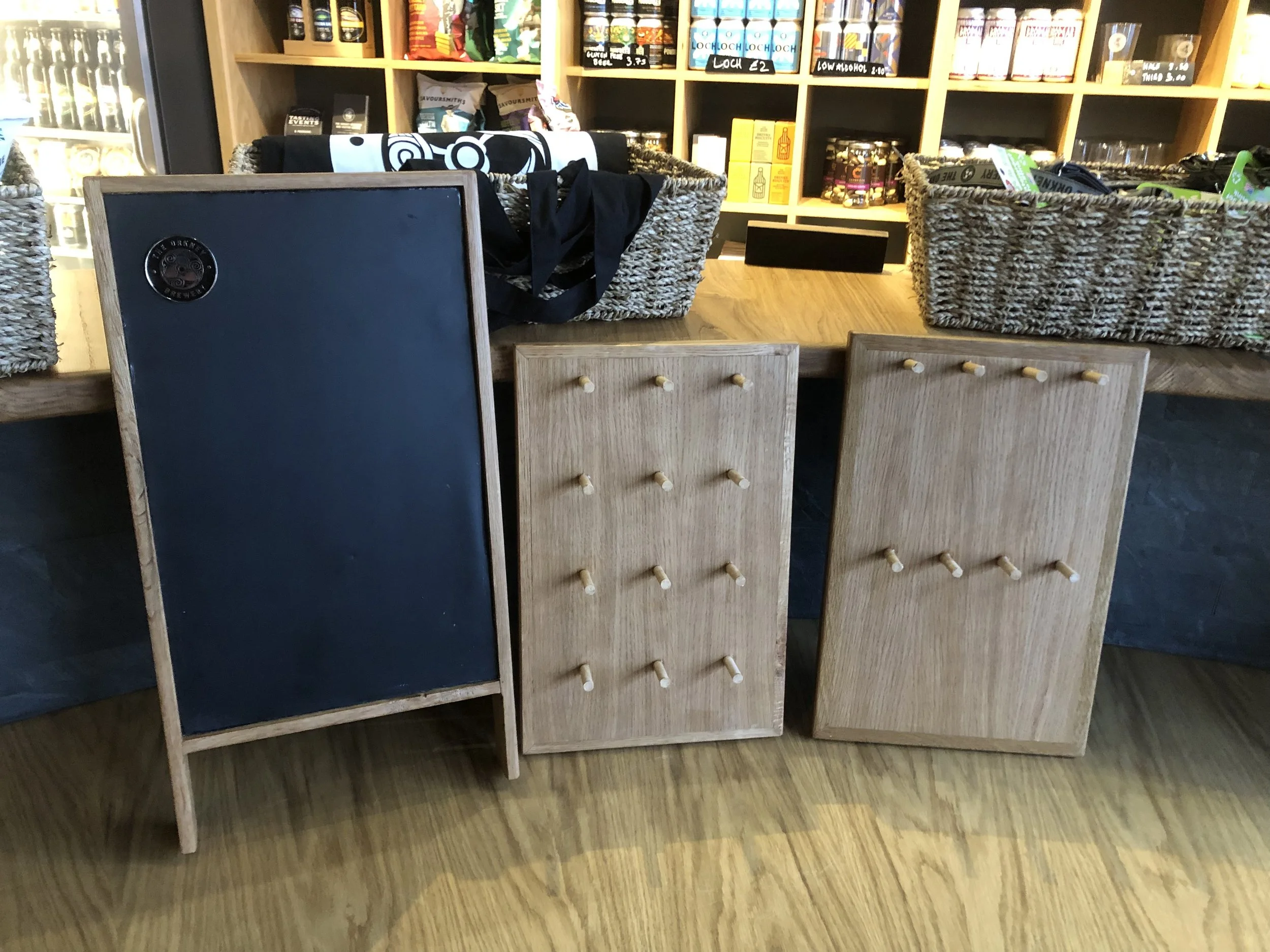 Three wooden boards, two with many small knobs, are propped against a table in a store. One board has a black chalkboard surface, and the other two have a natural wood finish with knobs. Wicker baskets and shelves filled with various products are on the table and shelves in the background.