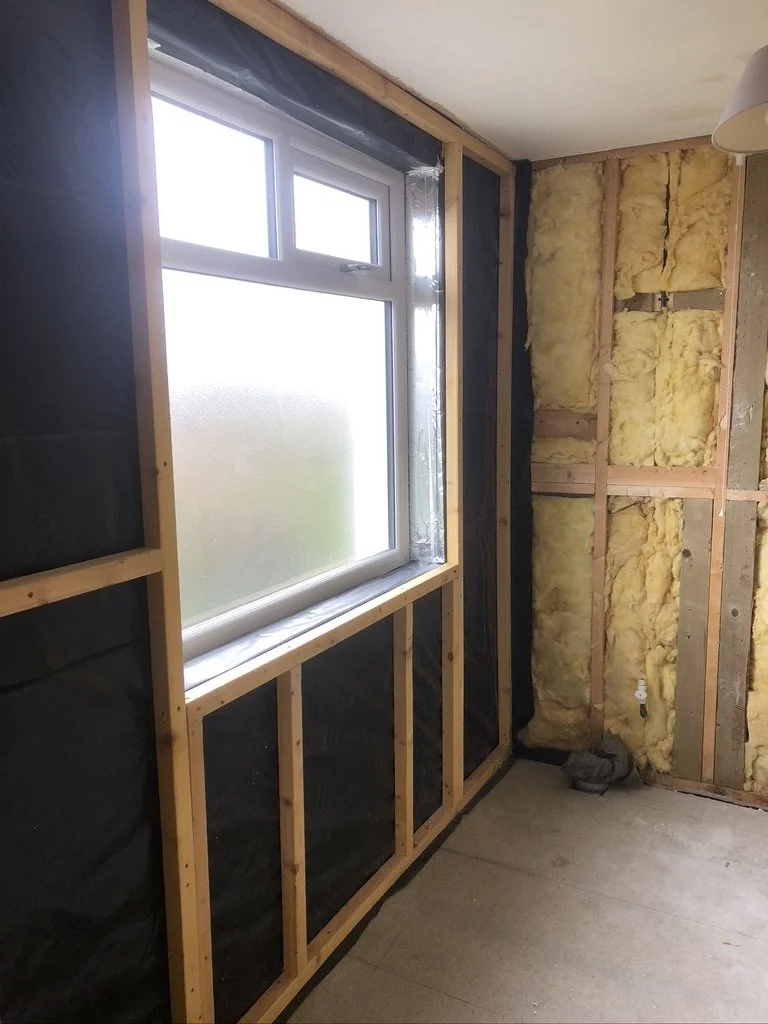 Interior of a room under renovation with a window, yellow insulation, and exposed wooden framing.