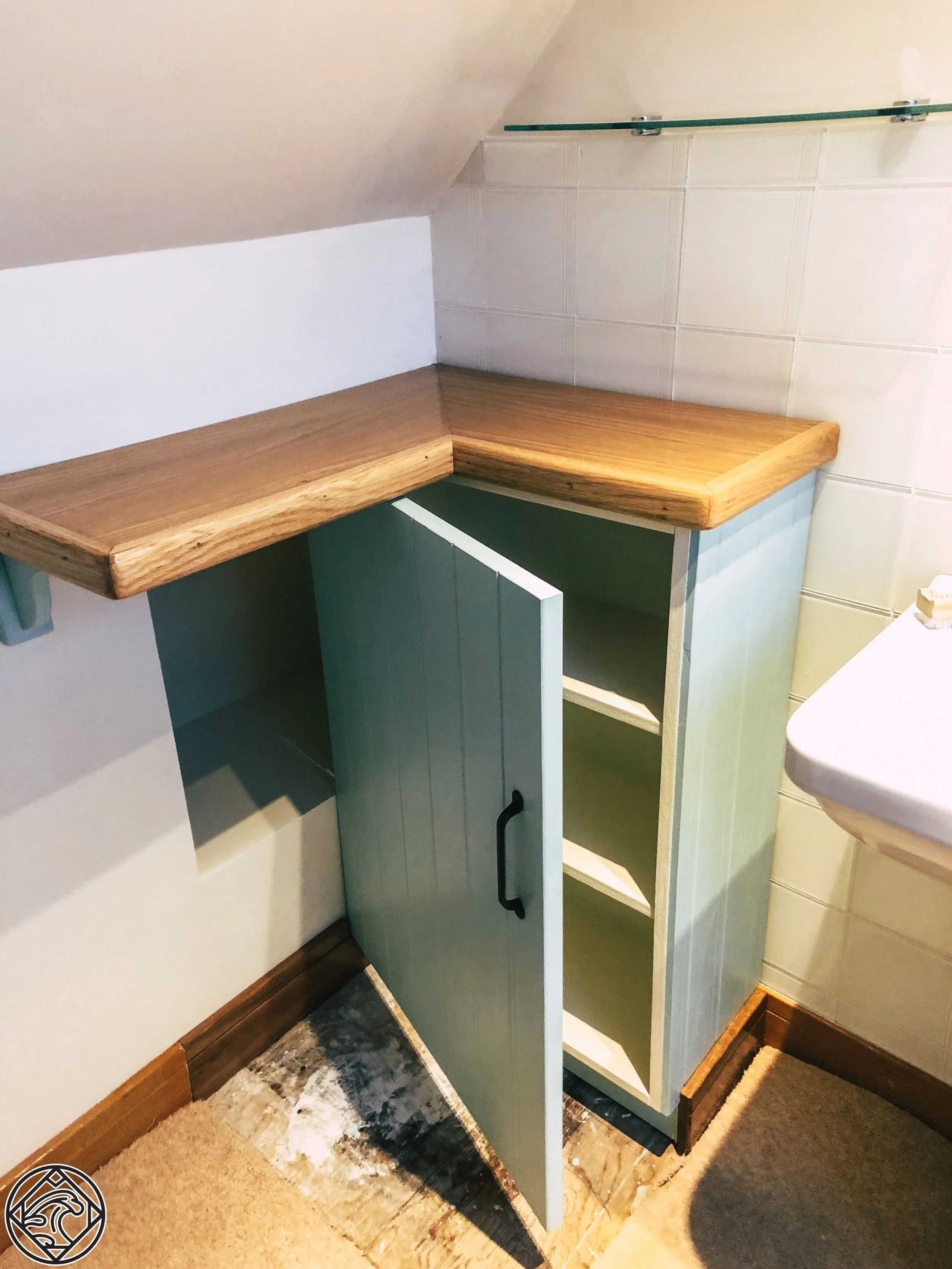 A small corner cabinet with a black handle and open shelves, topped with a wooden countertop in a room with white tiled walls and a beige floor.