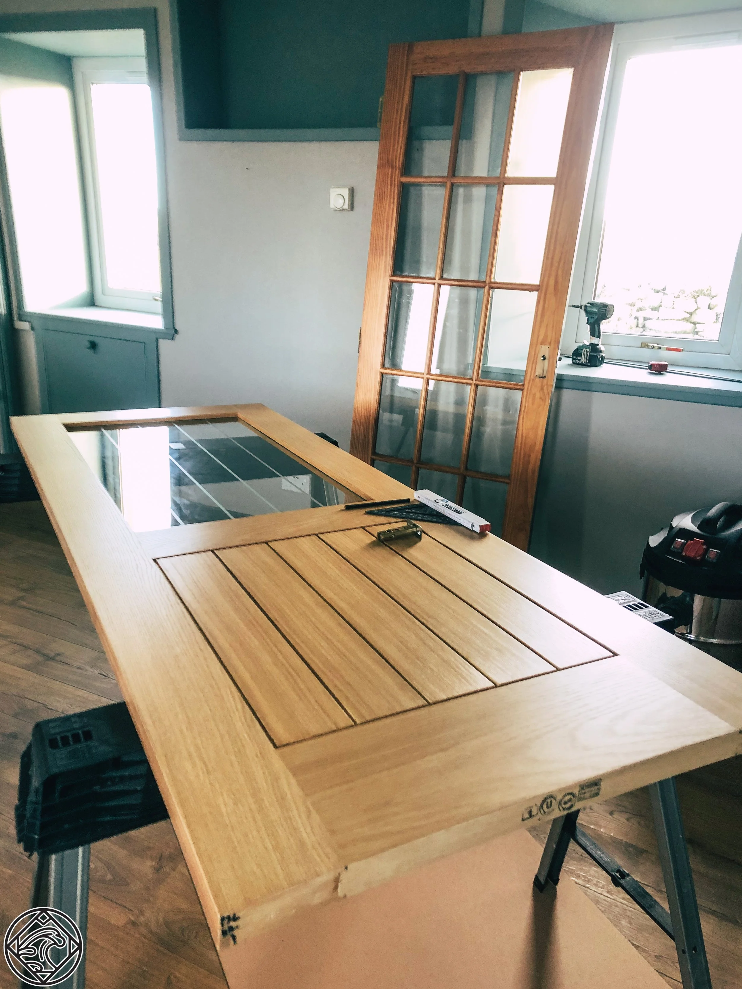Interior of a room with a wooden table, a window, and a glass-paneled wooden door leaning against the wall.