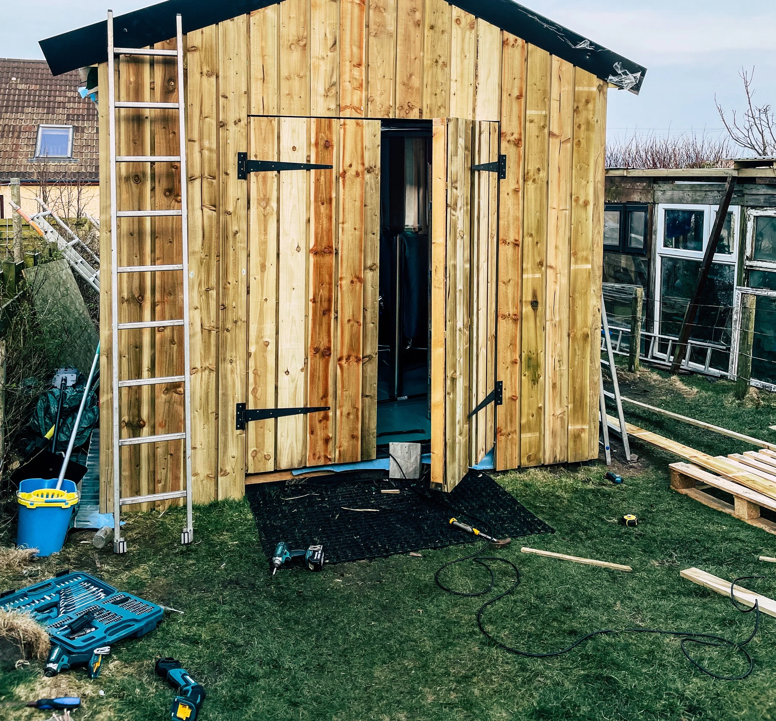 A wooden shed under construction with an open door, a ladder leaning against it.