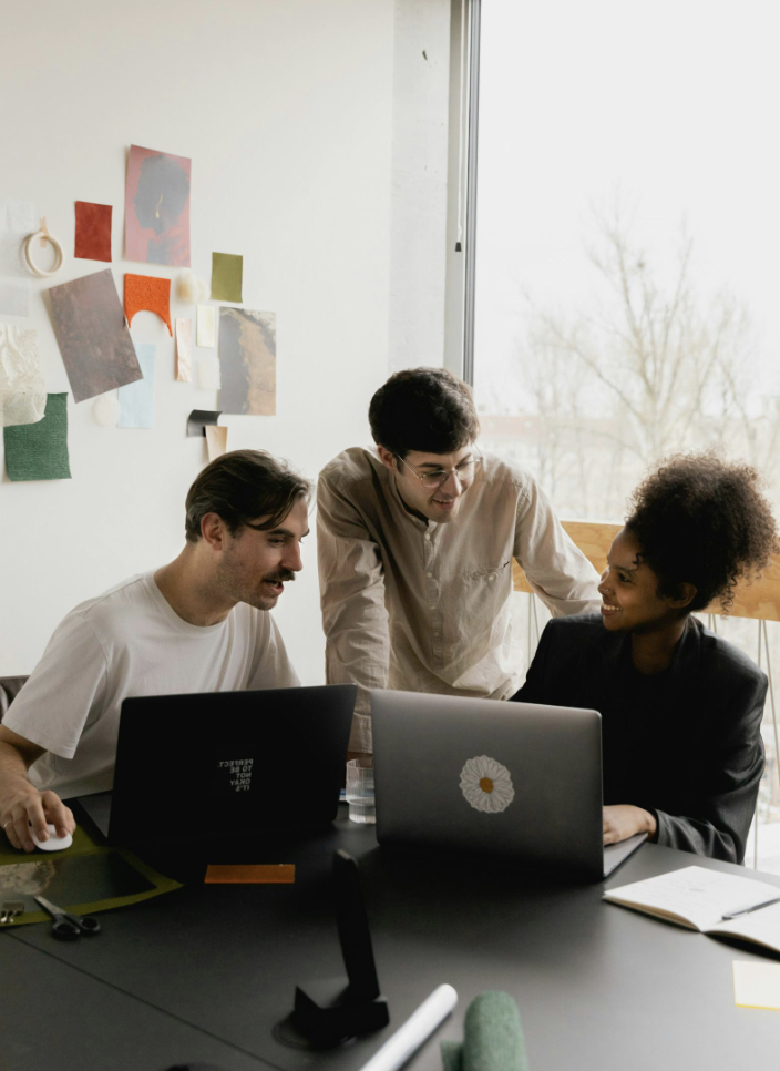 Drei Personen arbeiten gemeinsam an Laptops in einem hell erleuchteten Raum mit einer großen Fensterfront, hinter der Bäume zu sehen sind.