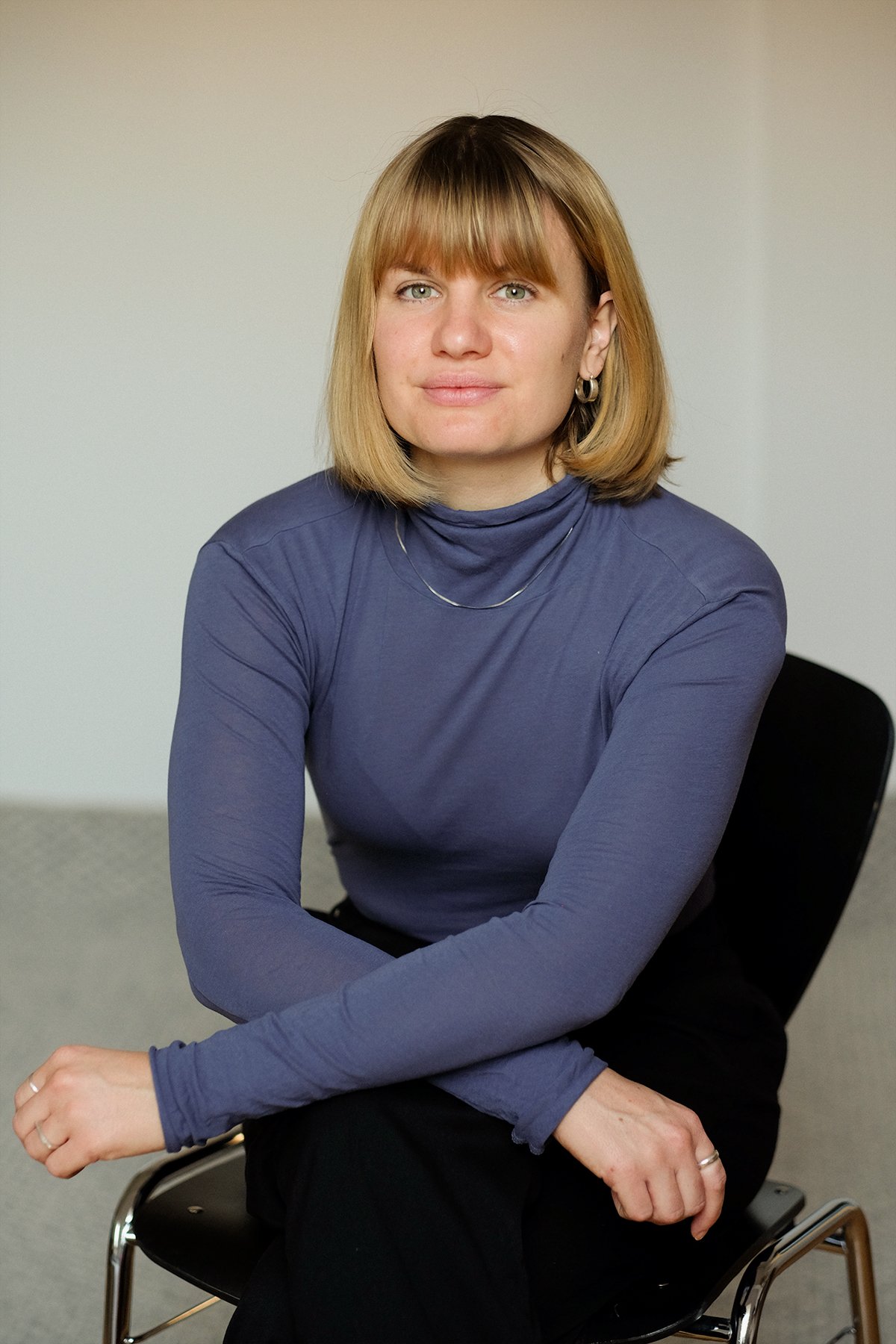 Porträt einer Frau mit blonden Haaren, blauen Augen, in einem blauen Rollkragenpullover, sitzt auf einem schwarzen Stuhl in einem Raum mit neutralem Hintergrund.