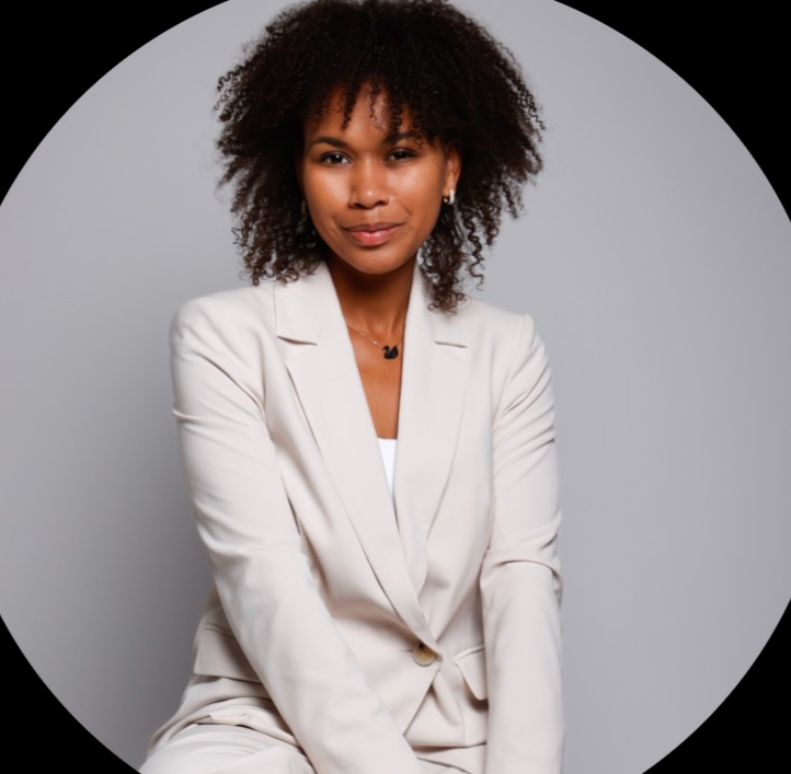 Eine junge Frau mit lockigem Haar, die einen weißen Business-Anzug trägt, vor einem grauen Hintergrund.