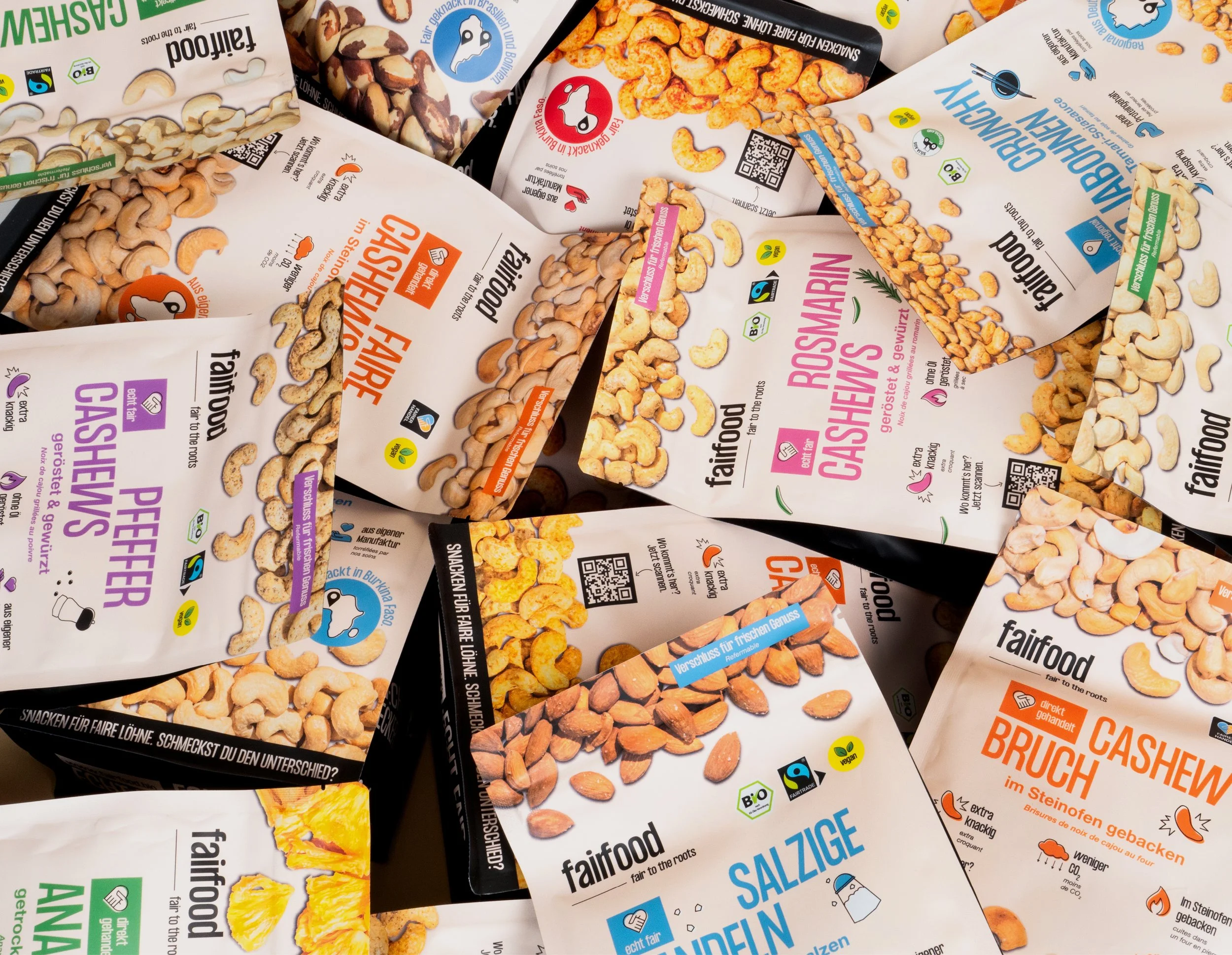 Mehrere Tüten mit fairtrade Cashewkernen, Cashew-Peffer, Cashewbruch und andere Snacks, alle mit bunten Aufklebern und QR-Codes auf weißem Hintergrund.