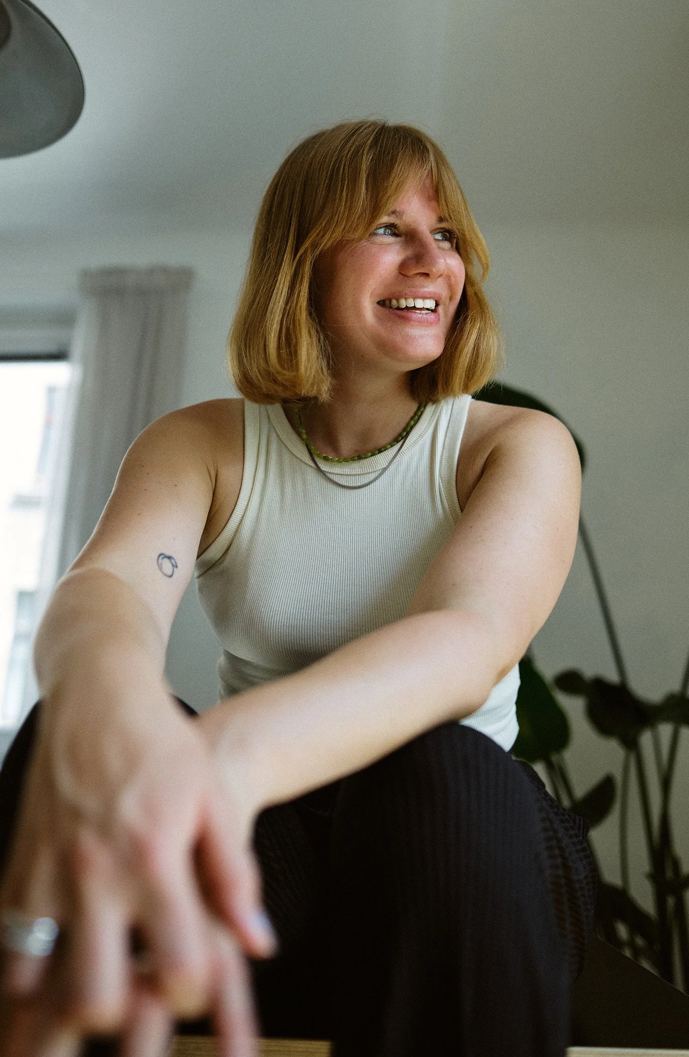 Junge Frau mit blondem Haar, lachend, Blick nach rechts, sitzend in einem Raum mit Pflanzen im Hintergrund, trägt eine weiße Top, Schmuck und einen Tattoo auf dem Arm.