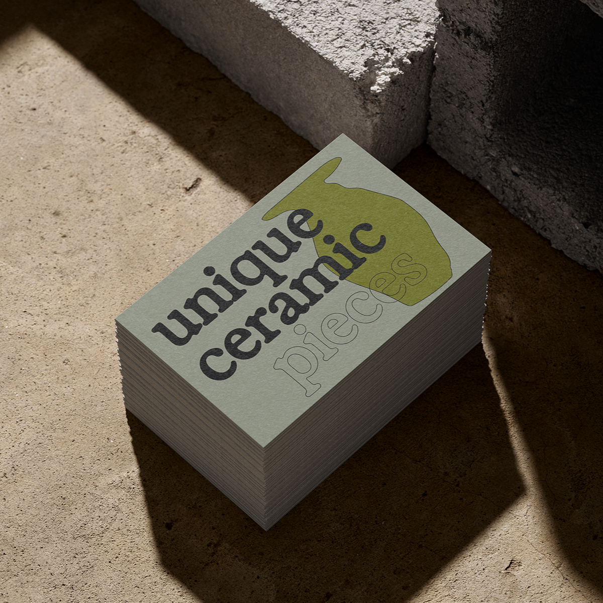 Ein Stapel Flyer oder Visitenkarten mit dem Logo und Namen "unique ceramics piec" auf einer sandigen Oberfläche, teilweise im Schatten.