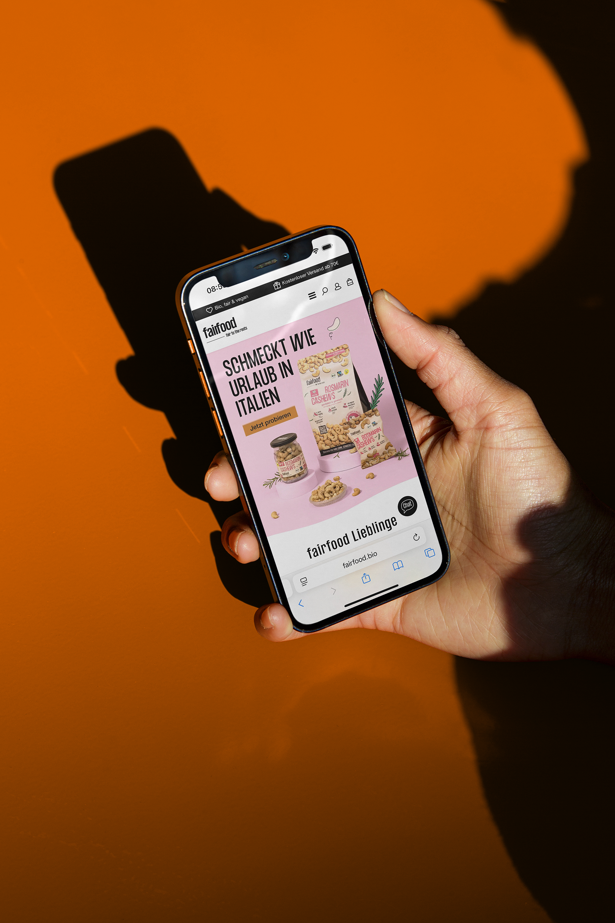 Hand hält Smartphone, das die Webseite von fairfood zeigt, mit einem Produktangebot für italienische Snacks auf pinkem Hintergrund.