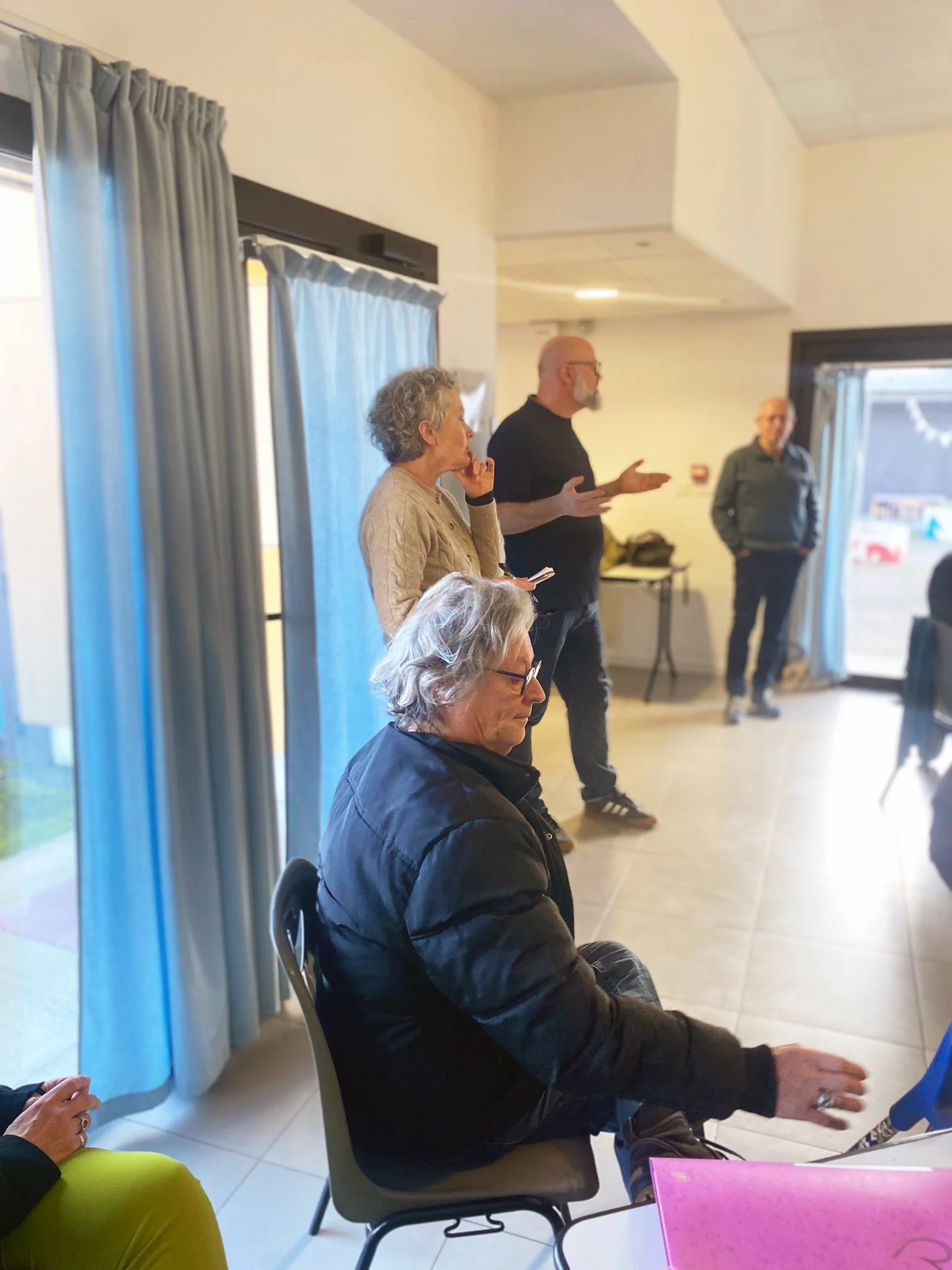 Groupe de quatre personnes dans une salle, une femmes âgée prenant la parole, trois autres assises ou debout qui écoutent, fenêtres avec rideaux, ambiance chaleureuse.