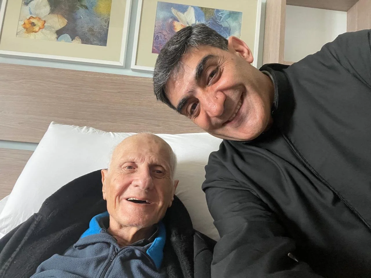A man and an elderly man smiling, taking a selfie together in a room with framed artwork on the wall behind them.